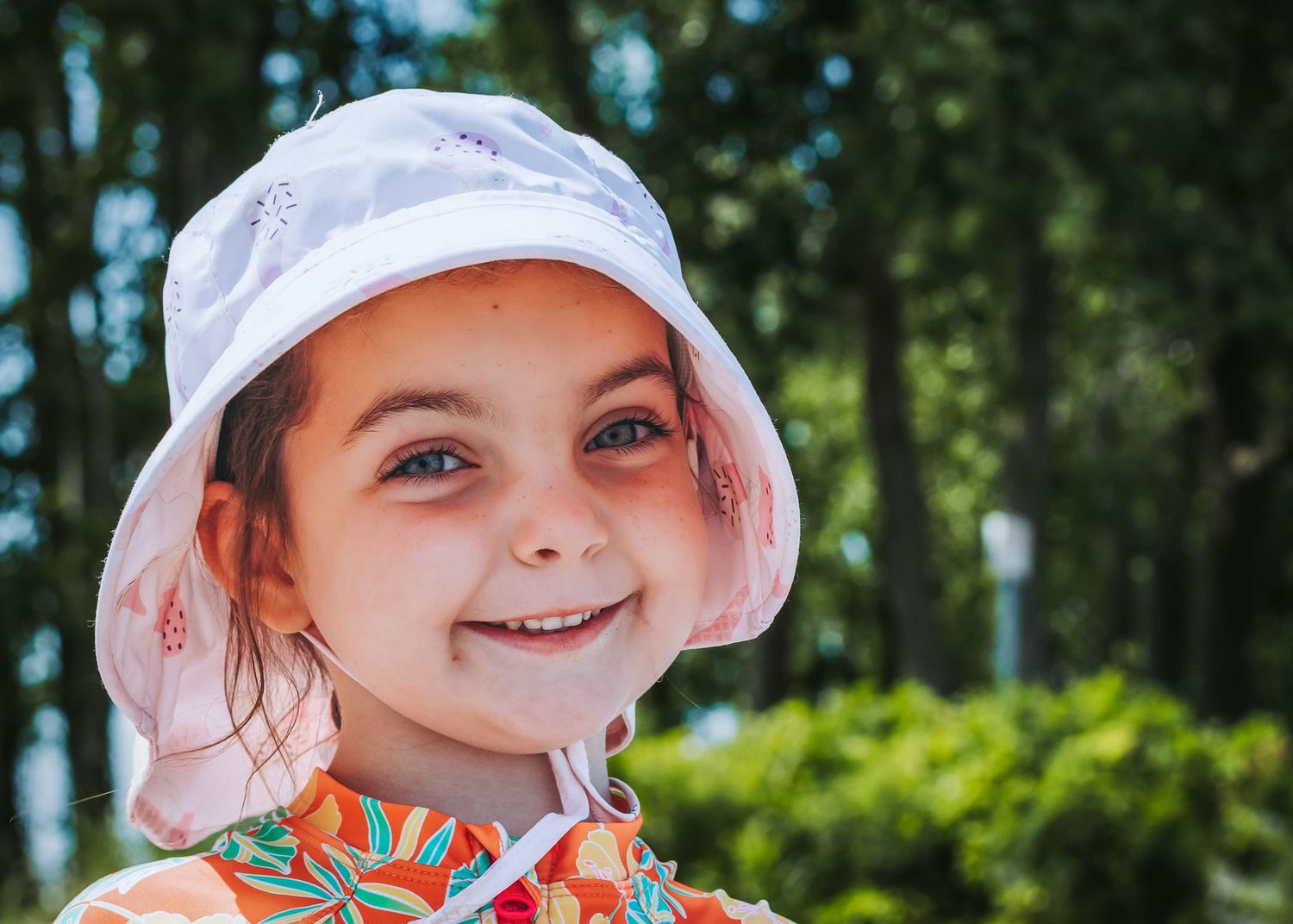 Young beachgoer in a white sun hat smiling against a backdrop of green trees on a sunny summer day.