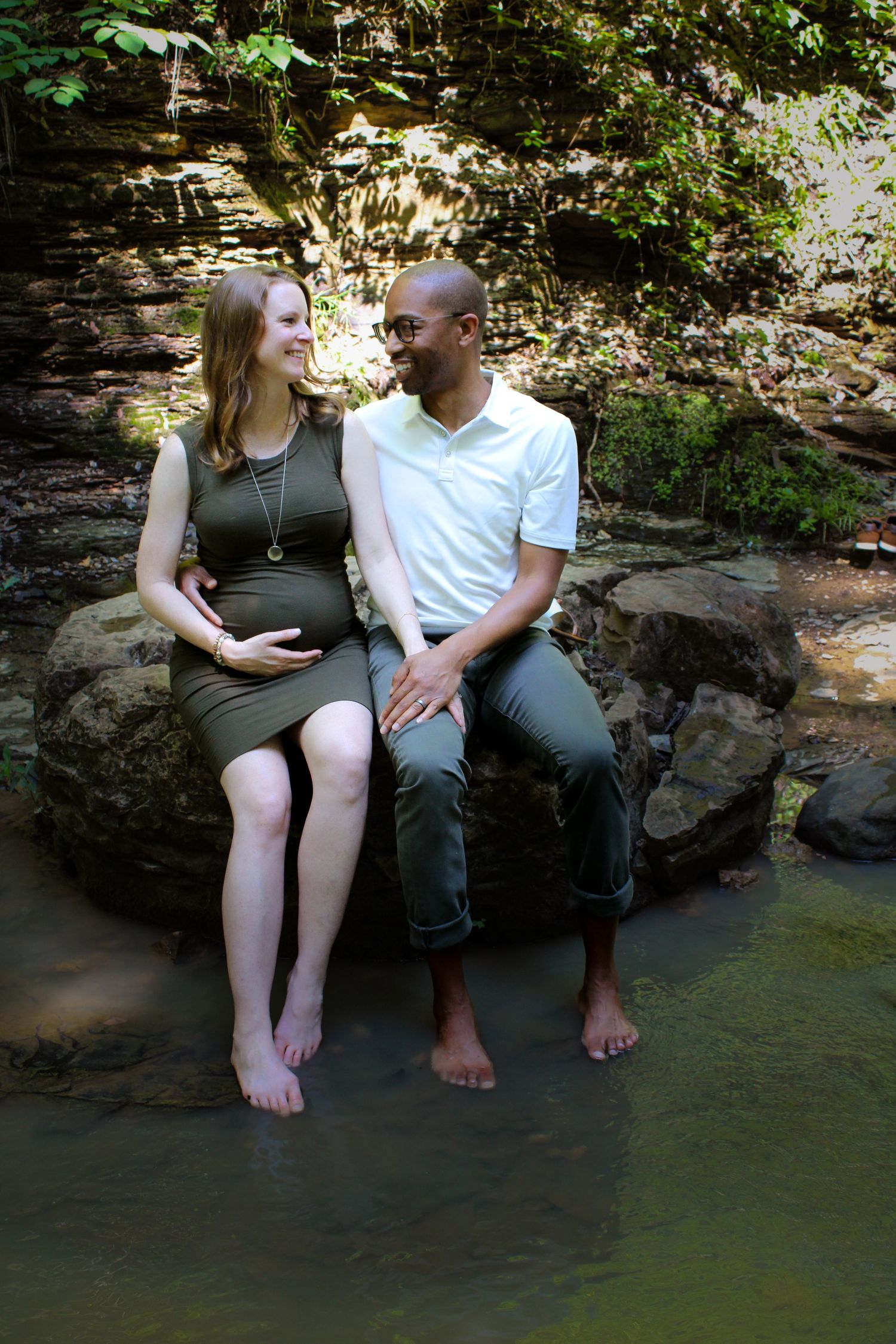 Couple enjoying a relaxing moment sitting on rocks by a peaceful stream on a sunny day.