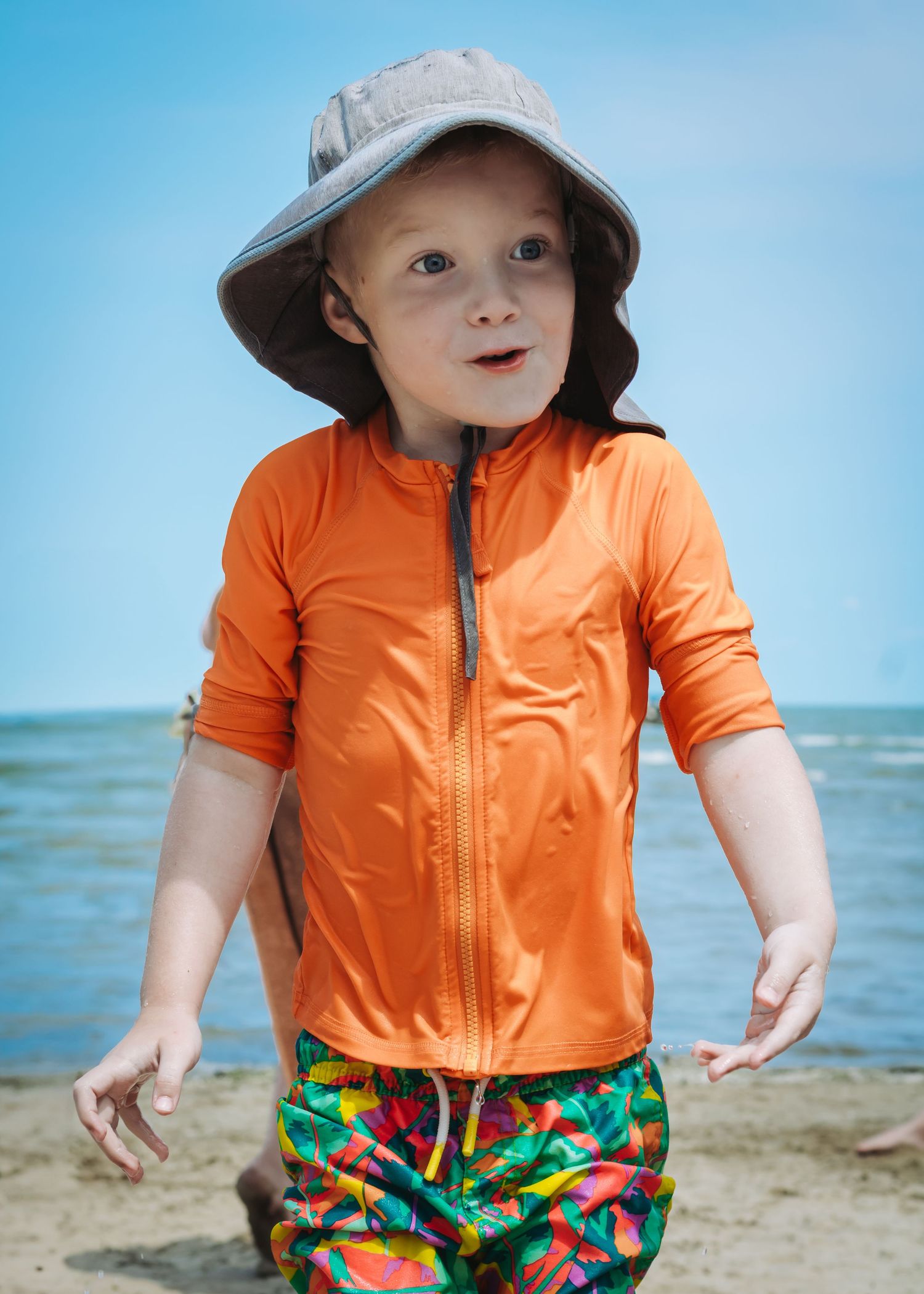 Small child playing on a sandy beach wearing bright orange jacket and colorful tropical print shorts by the ocean.