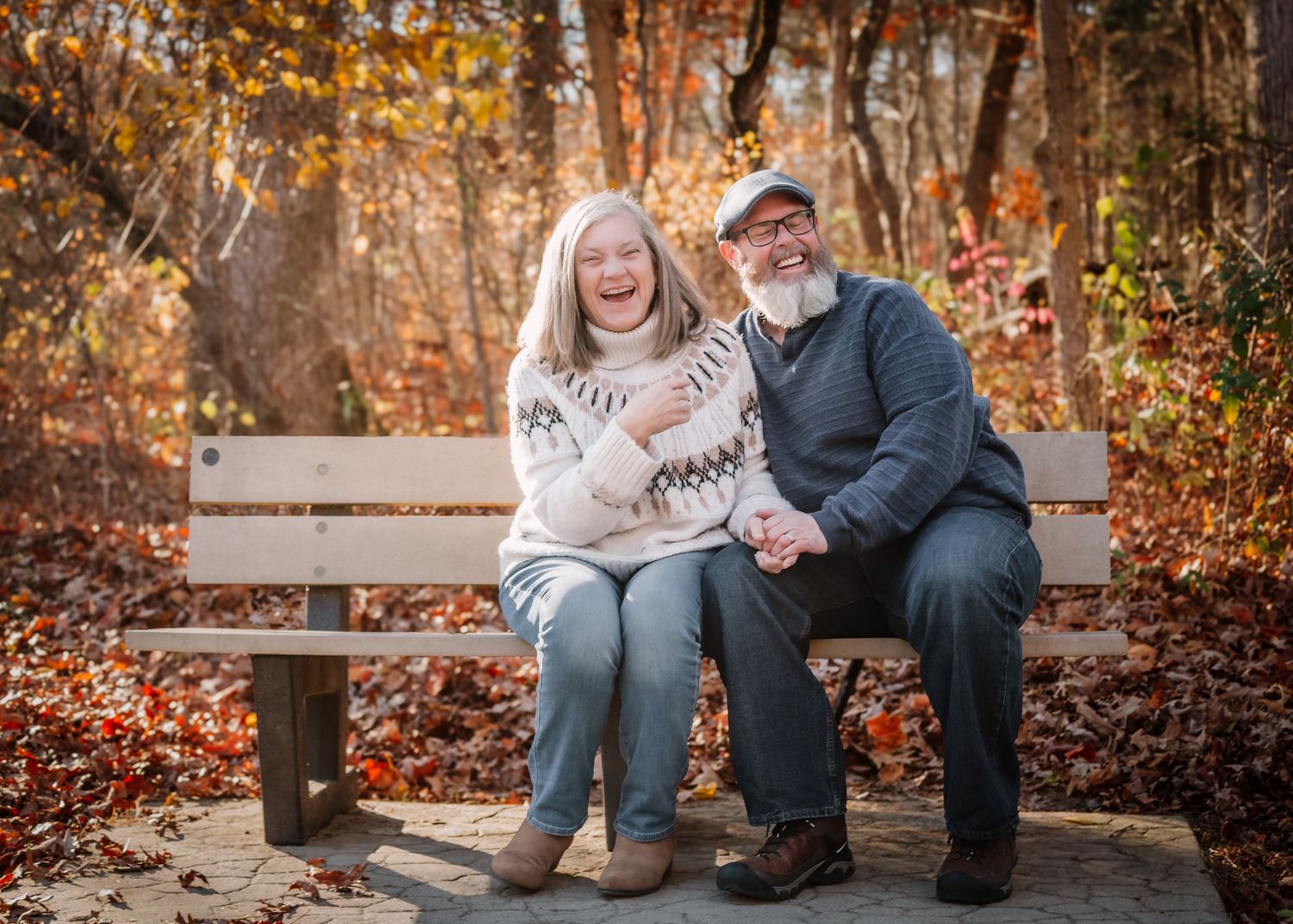 A couple sits on a wooden bench sharing a tender moment surrounded by autumn leaves in a park setting.