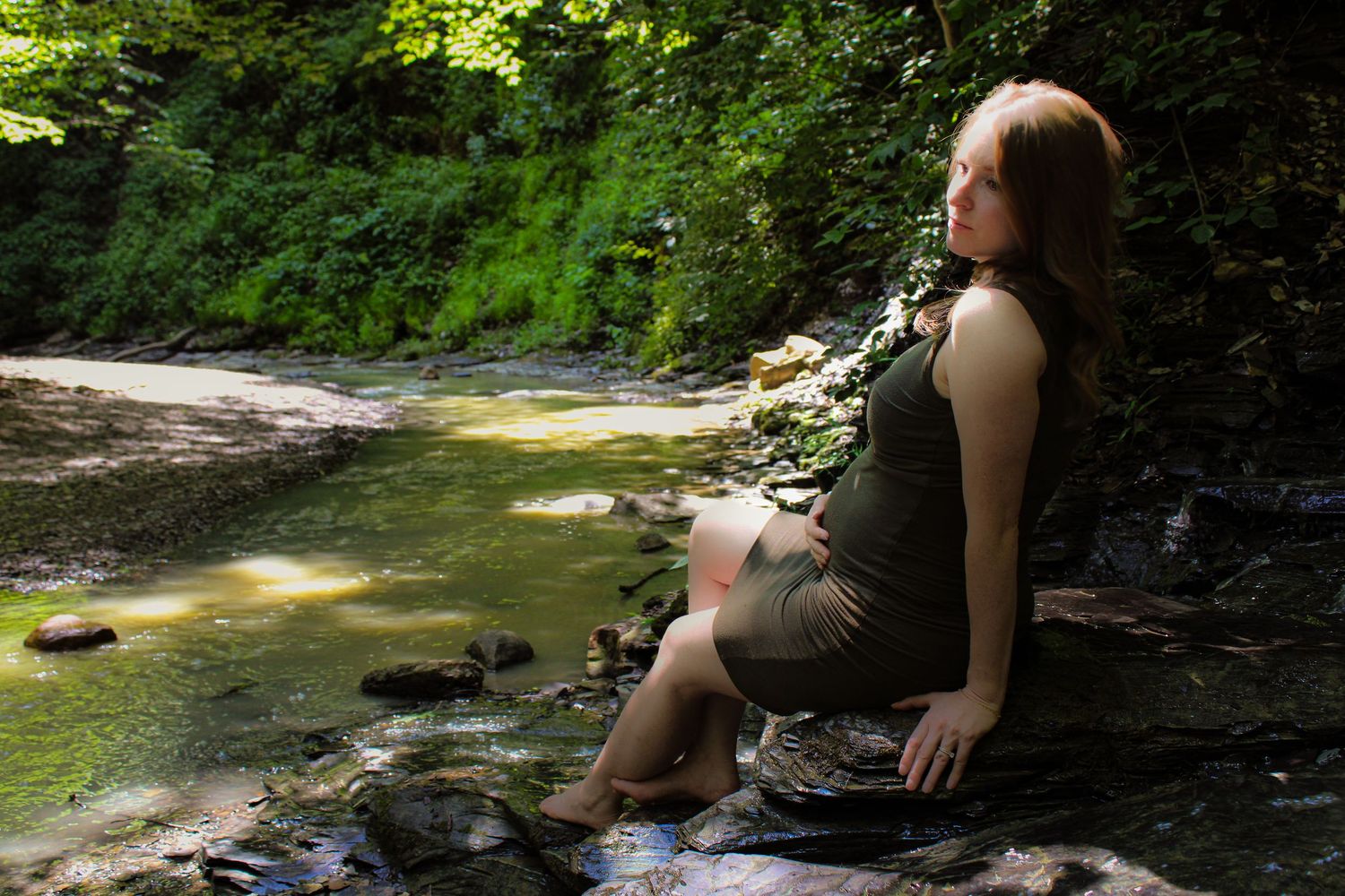 Person relaxes next to scenic forest stream with mossy rocks and lush greenery in summer afternoon light.
