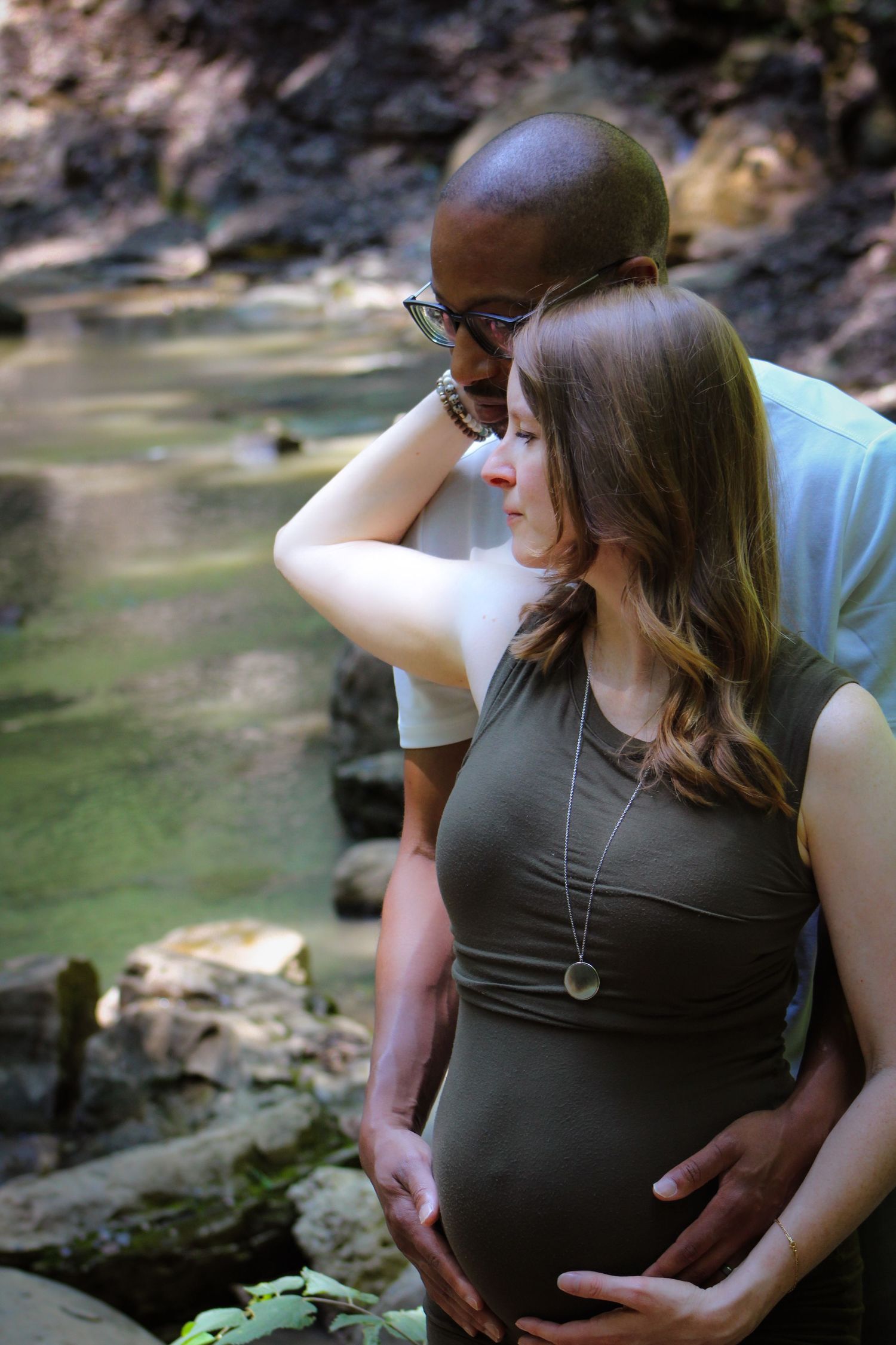Expecting couple embraces near a creek as they touch her pregnant belly in an intimate maternity photoshoot outdoors.