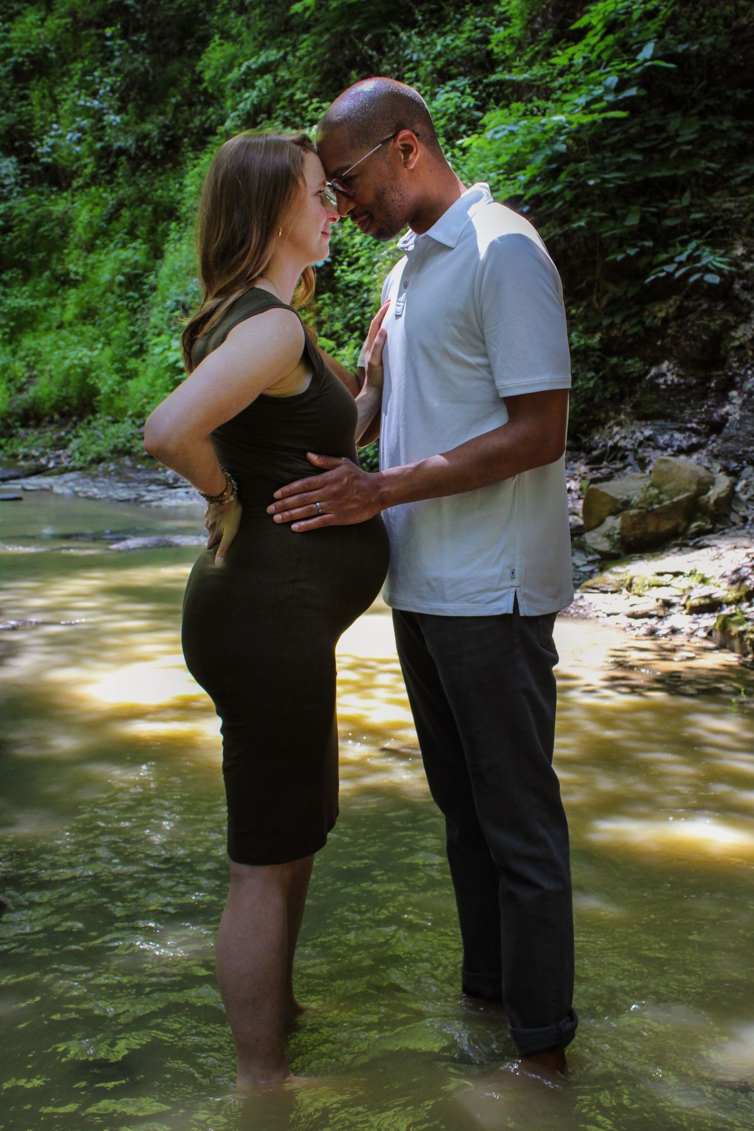 A romantic couple embraces while standing in a shallow creek with lush green foliage in the background.