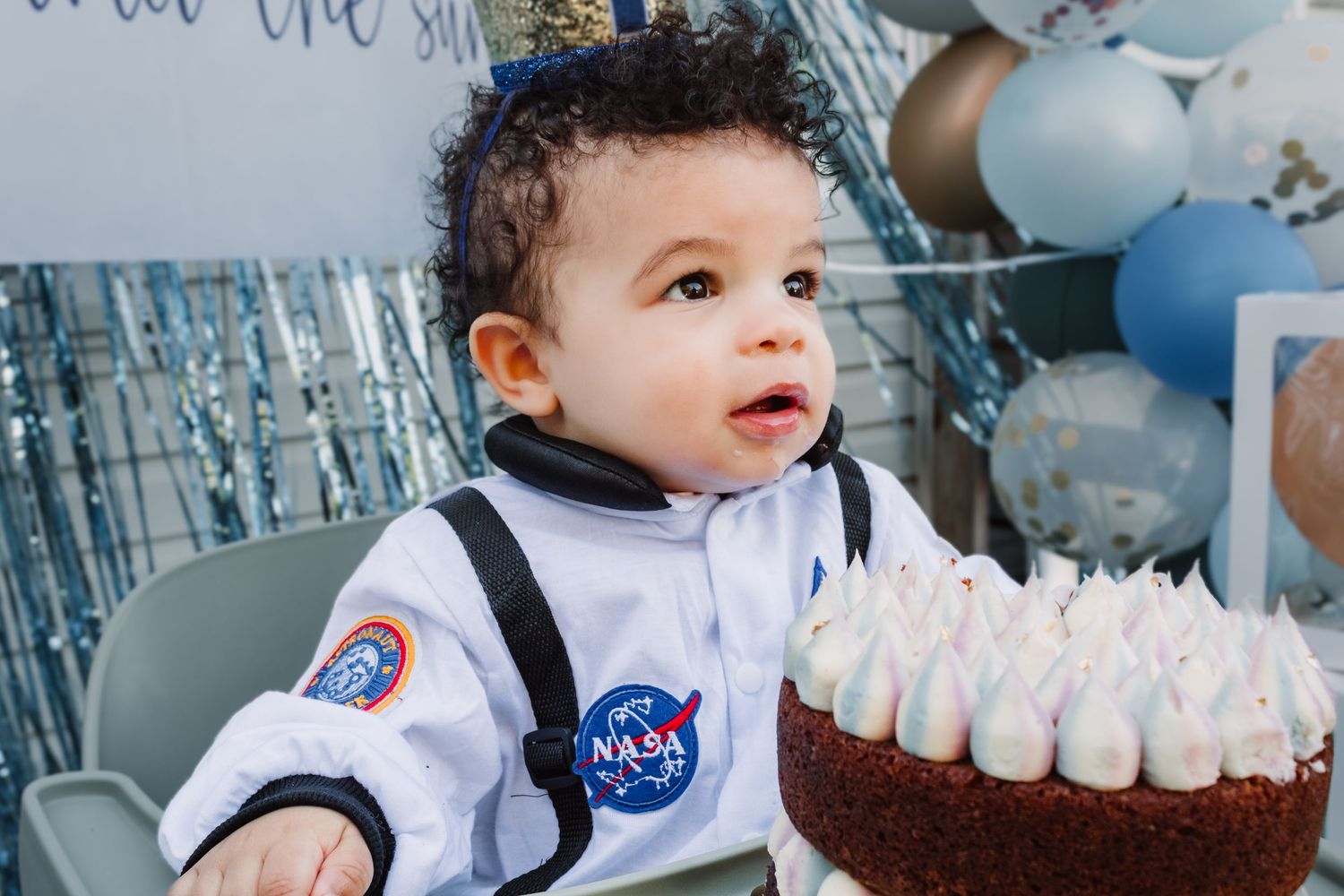 Child in NASA outfit sits in front of space-themed birthday cake and balloon decorations.