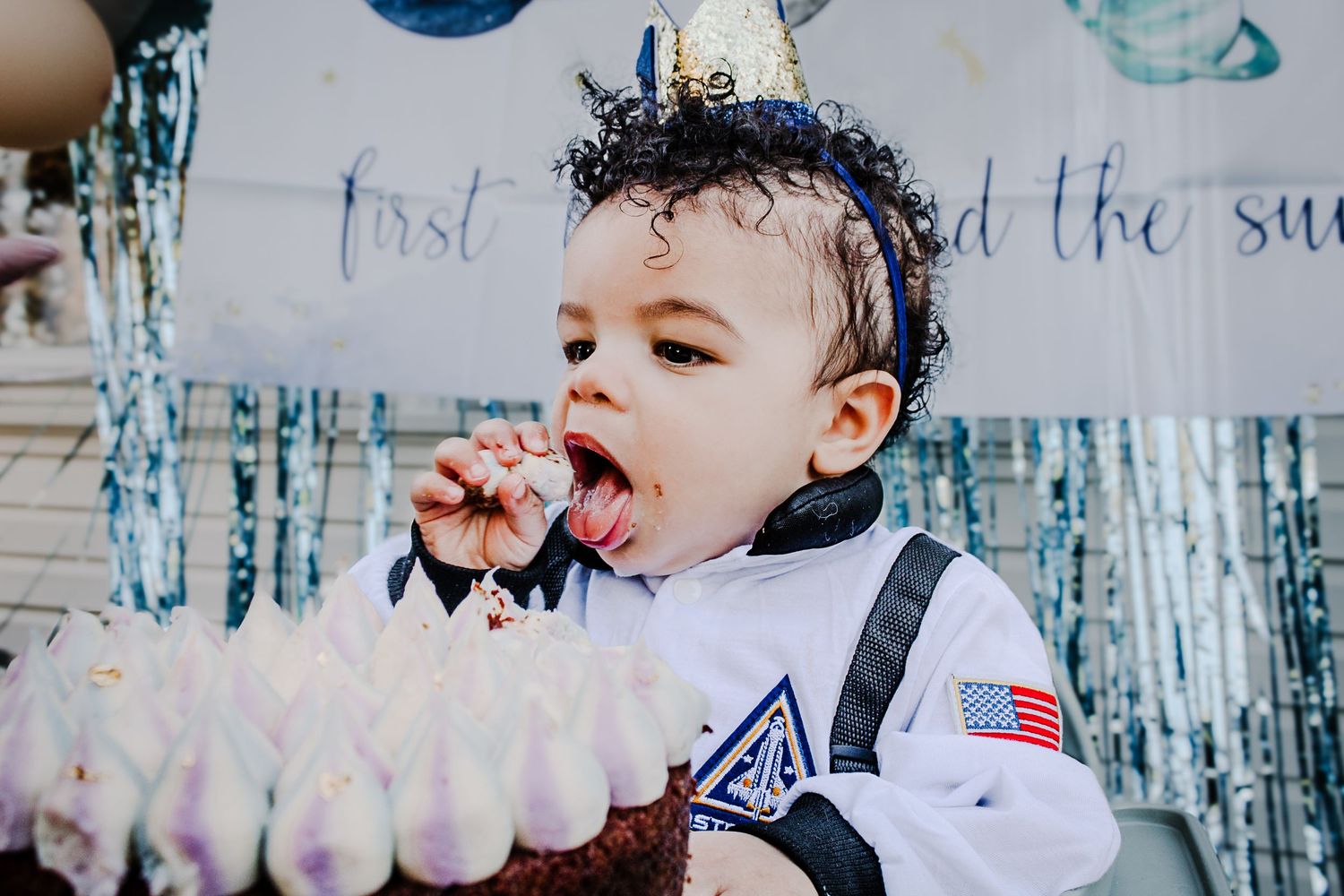 A happy baby wearing a white shirt and denim overalls plays while giggling near a festive shiny curtain backdrop.