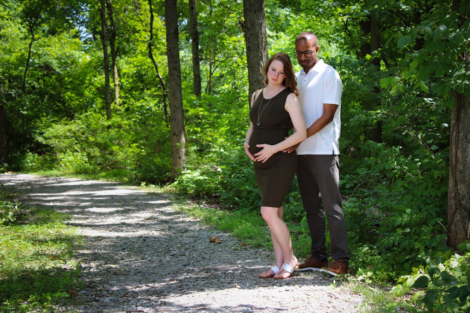 Couple walking together on a shaded woodland trail surrounded by lush green foliage on a sunny day.
