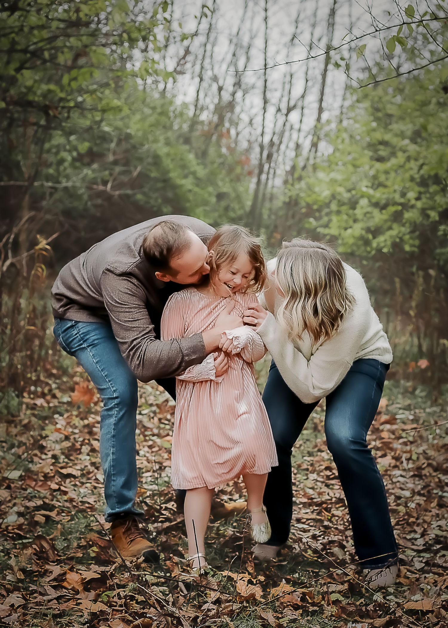 Joyful family moment captured in autumn forest with fallen leaves as parents share affectionate kisses with their child.