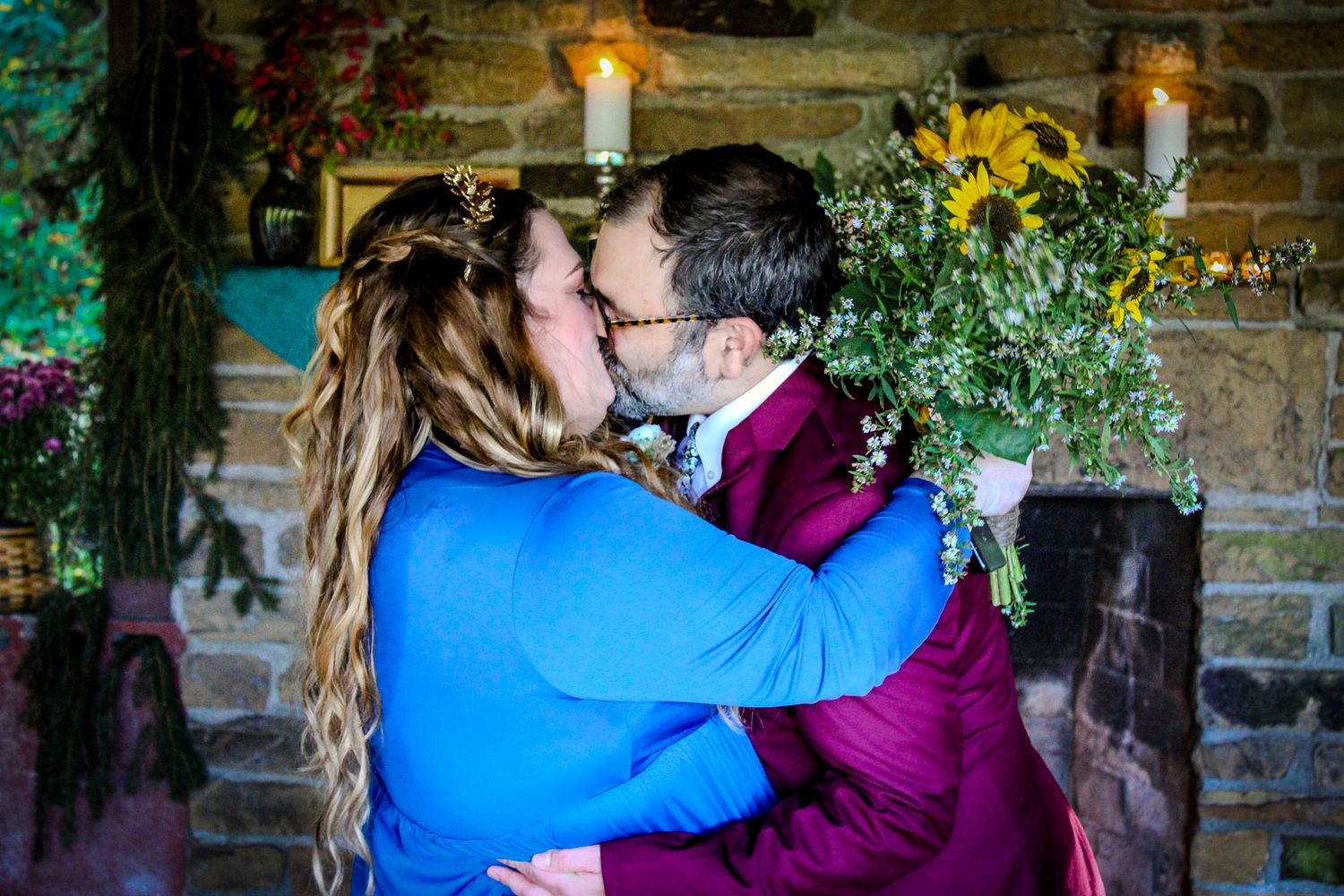 A romantic kiss between two people in bright blue and burgundy clothing against a rustic stone wall with glowing candles.
