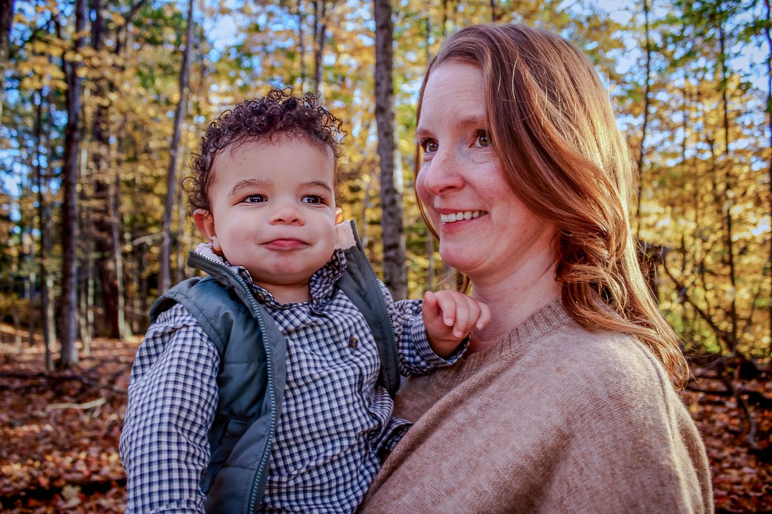A caring parent holds a toddler wearing a checkered shirt in a vibrant autumn forest setting.