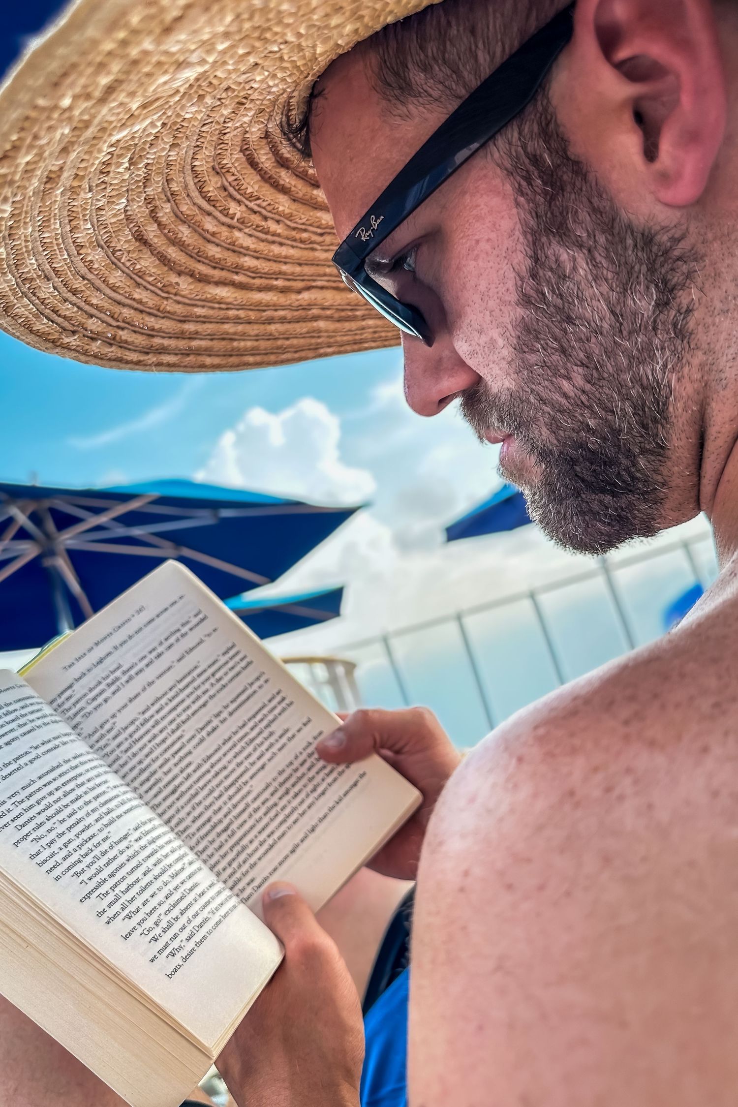 Person reading a book while relaxing under a straw hat on a sunny beach day.