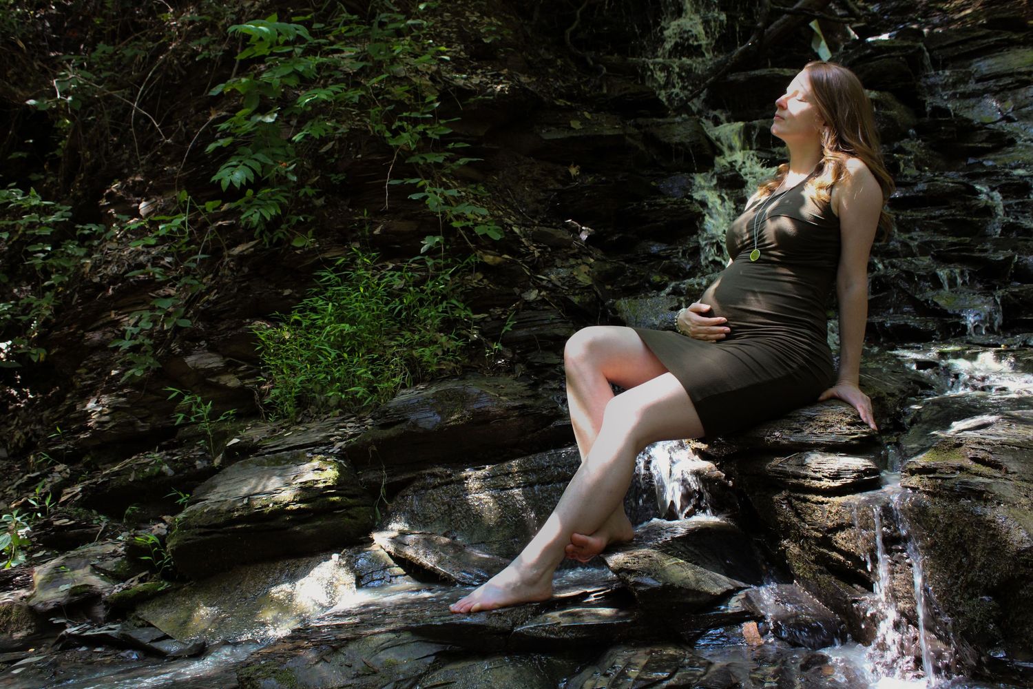 People relax and enjoy a sunny day by a cascading waterfall and rocky stream in a lush forest setting.