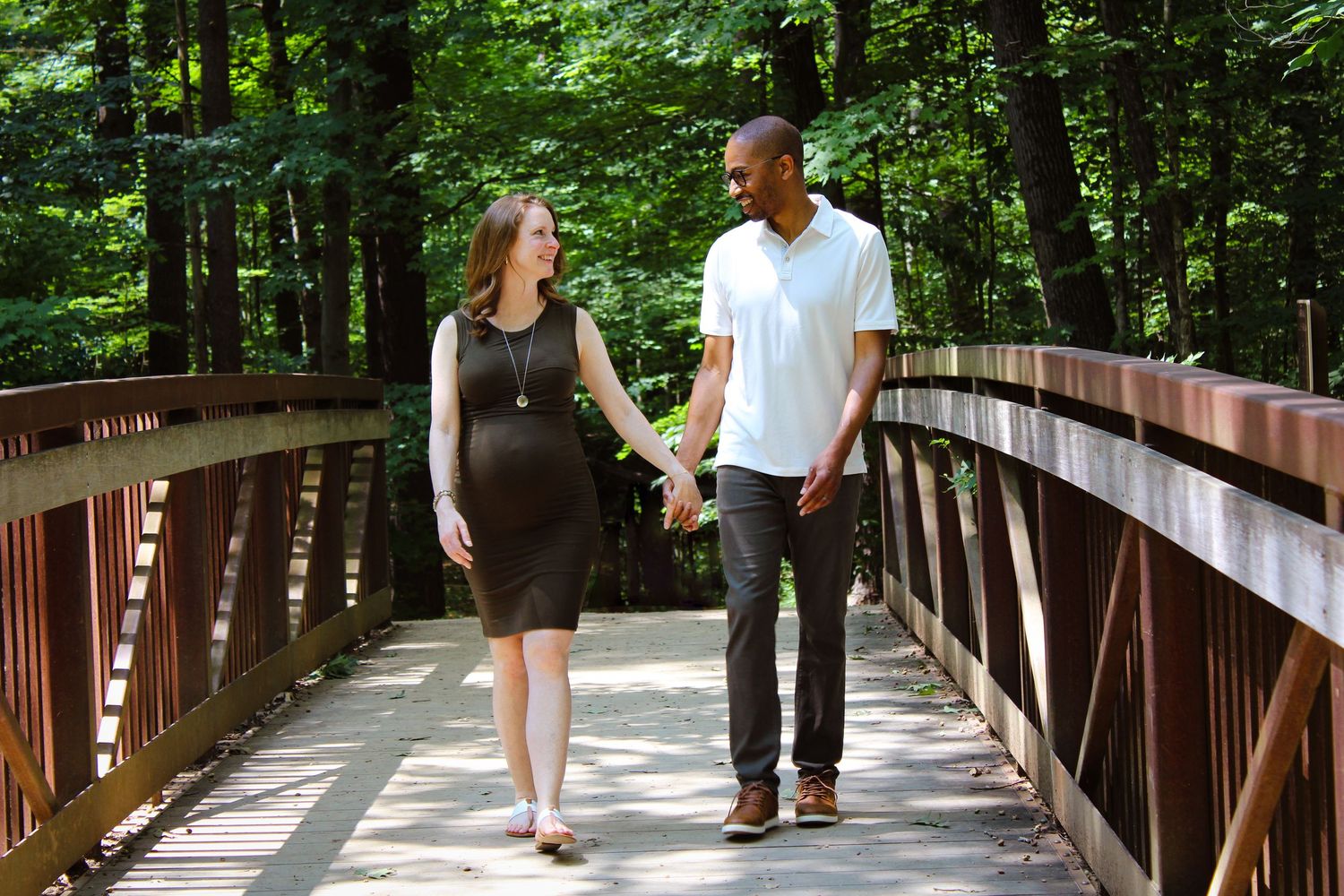 People stroll across a wooden bridge walkway surrounded by lush green forest on a sunny summer day.