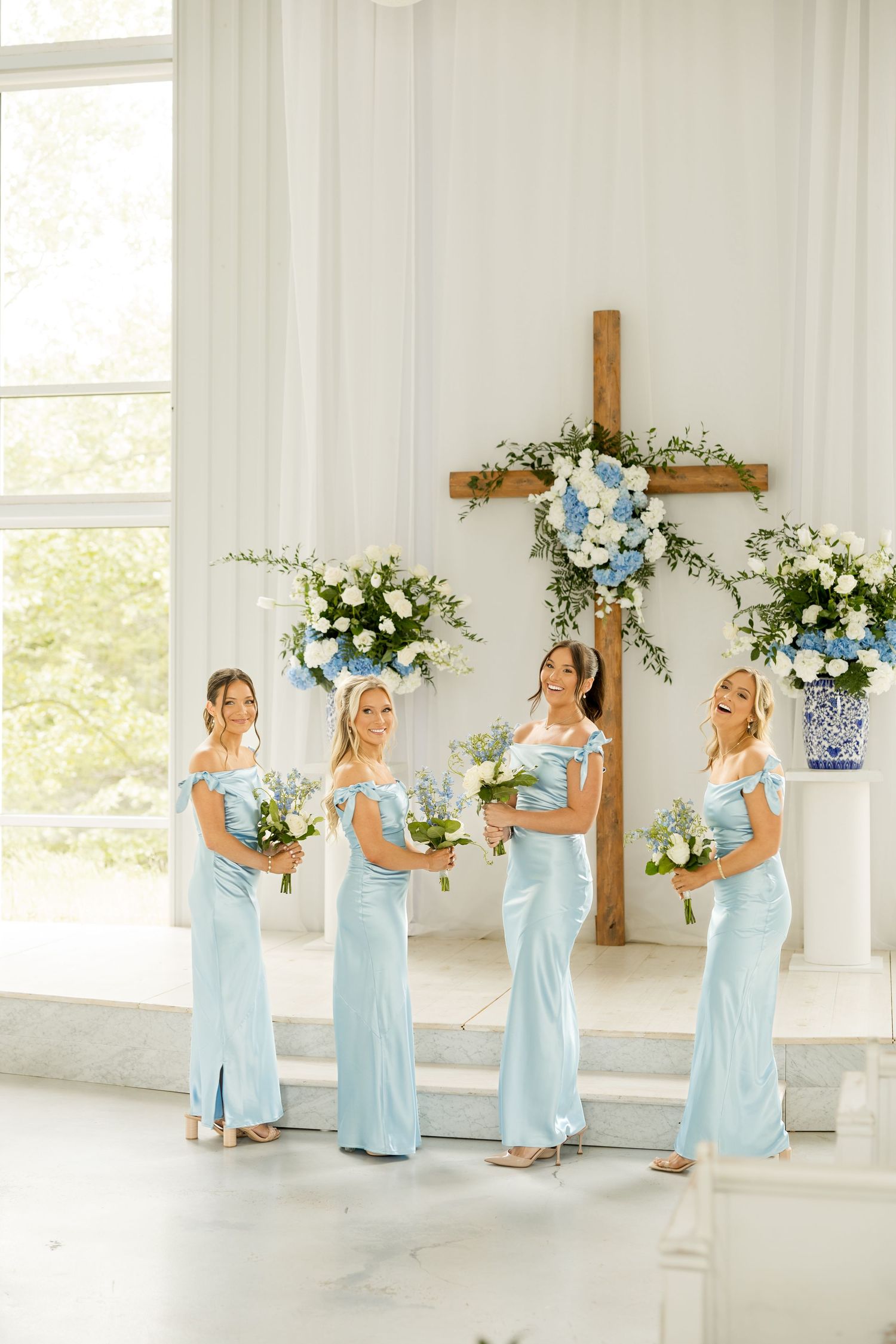 Bridesmaids in light blue dresses posing with bouquets in a bright white chapel. Cold Springs Events