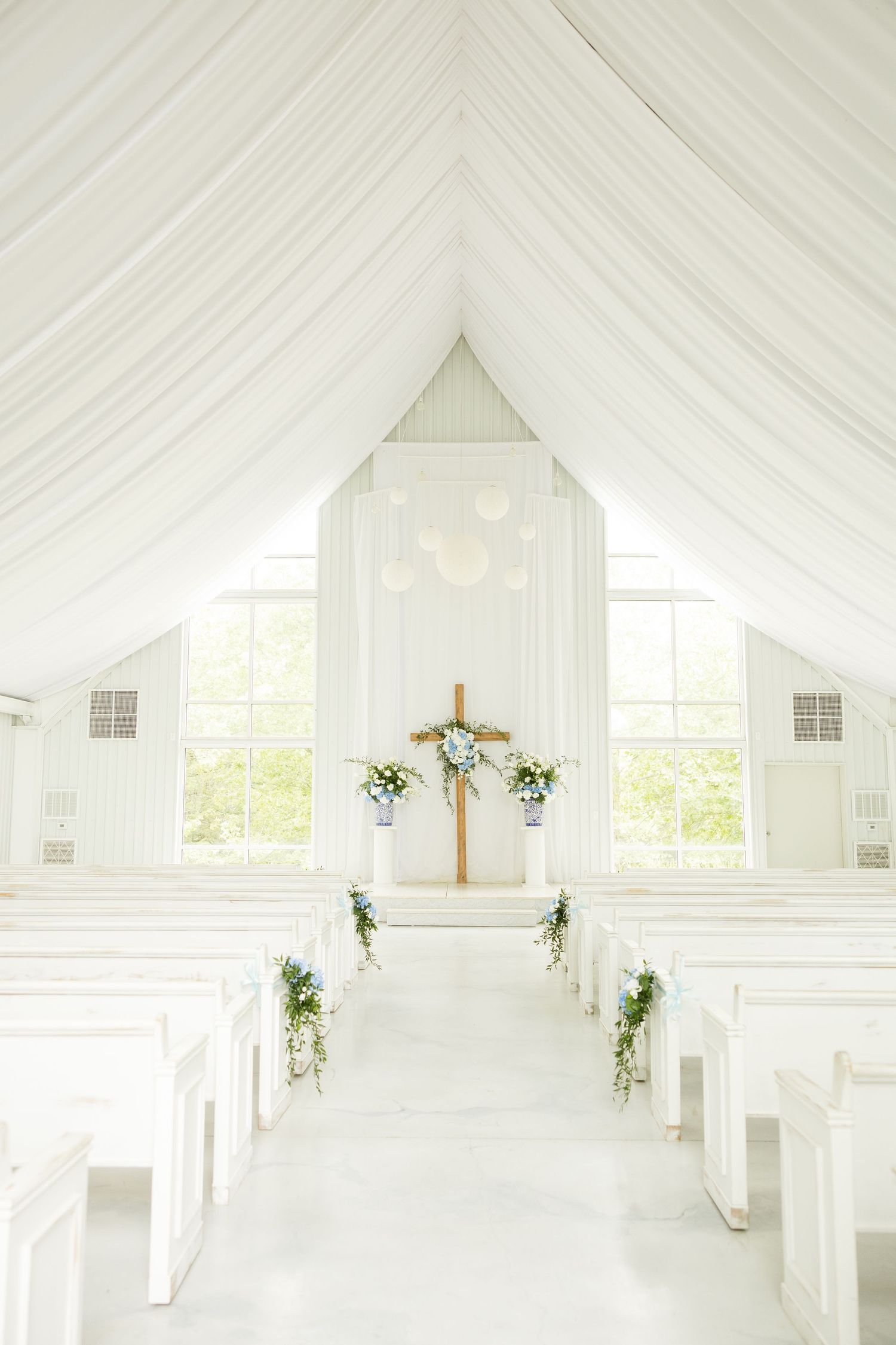 An all-white chapel interior with draped fabric ceiling and wooden cross at the altar.Cold Springs Events.