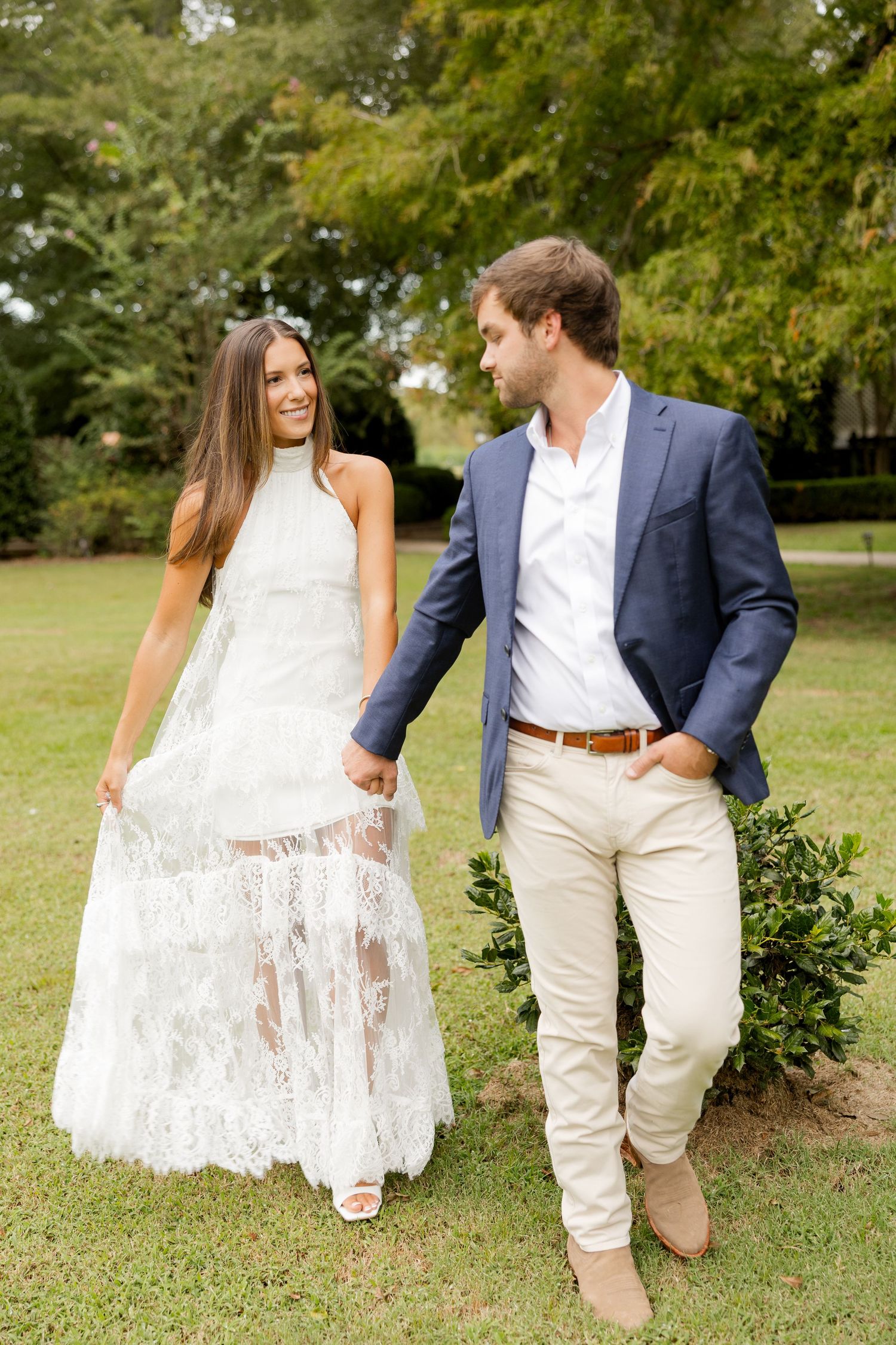 A couple poses together outdoors in formal attire, with the bride in a white lace dress and groom in a navy blazer.