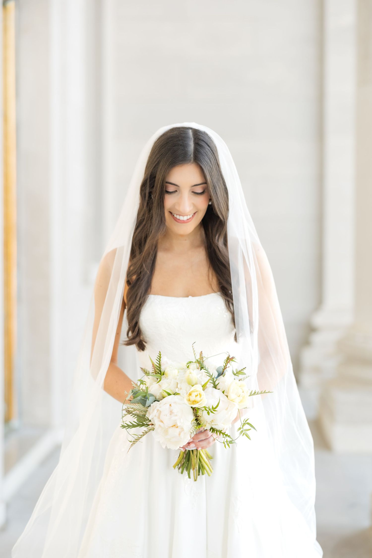 A bride in a strapless white gown smiles while holding a delicate bouquet of cream and white flowers.