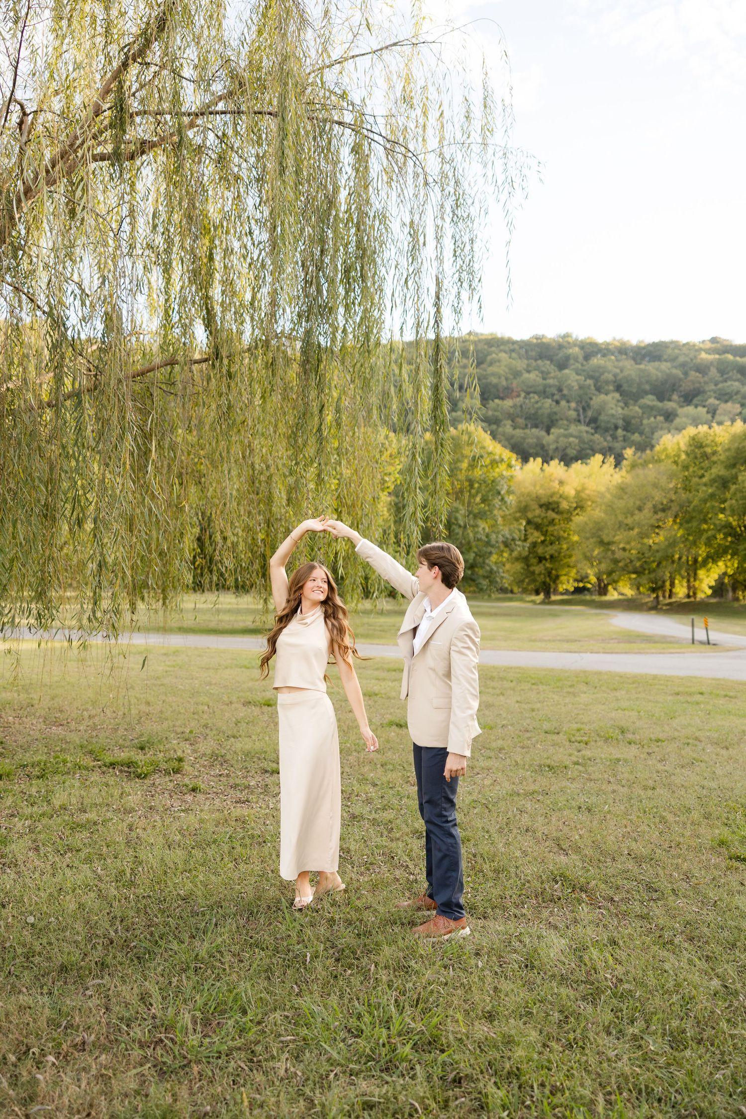 A couple shares a tender moment under a weeping willow tree in a serene outdoor setting during golden hour.