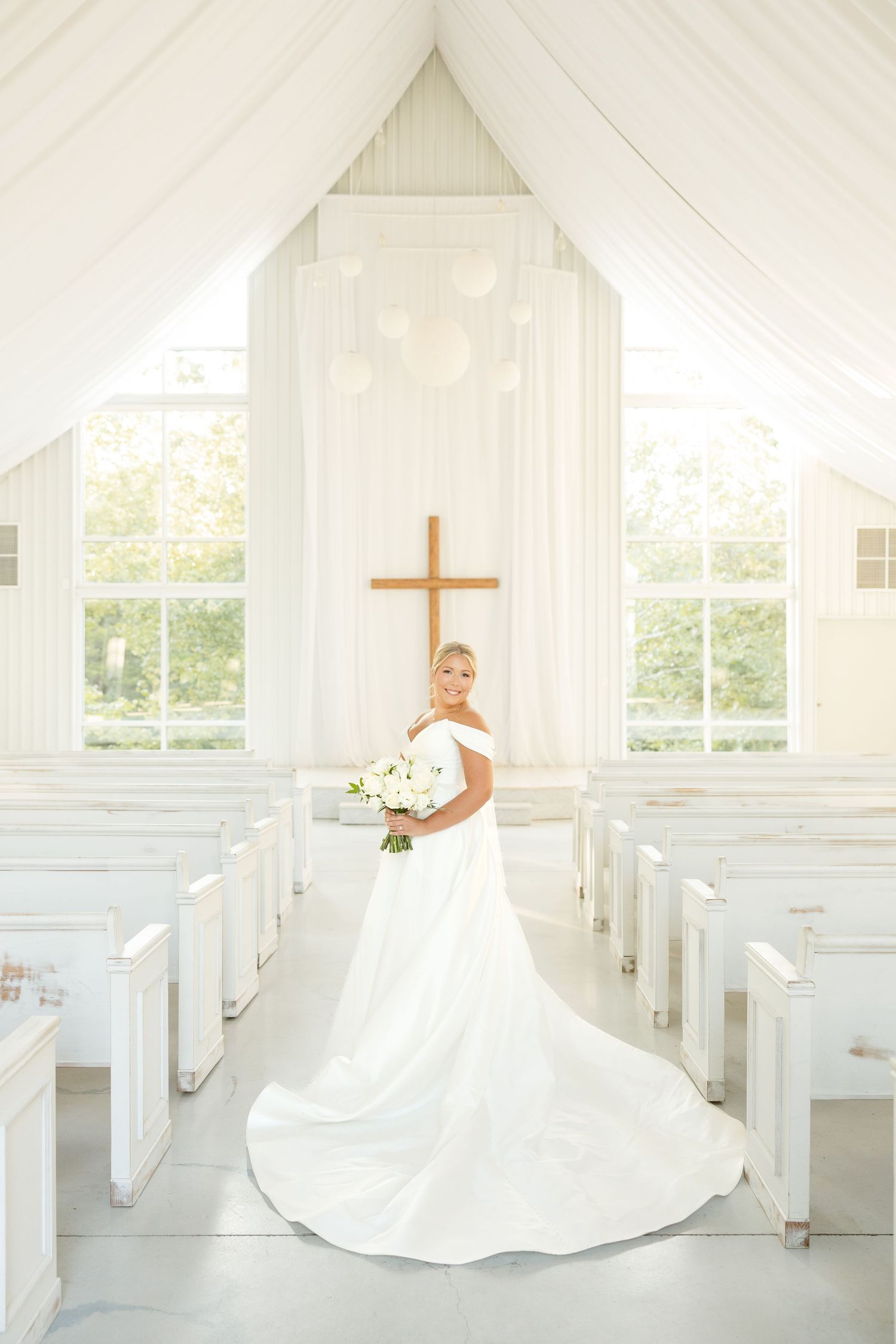 A bright white chapel interior features elegant draping and pews illuminated by natural window light.