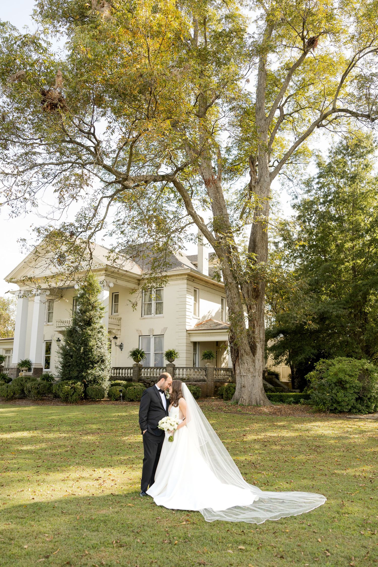 Wedding couple poses on lawn in front of white mansion on autumn day. Marlgsate Plantation