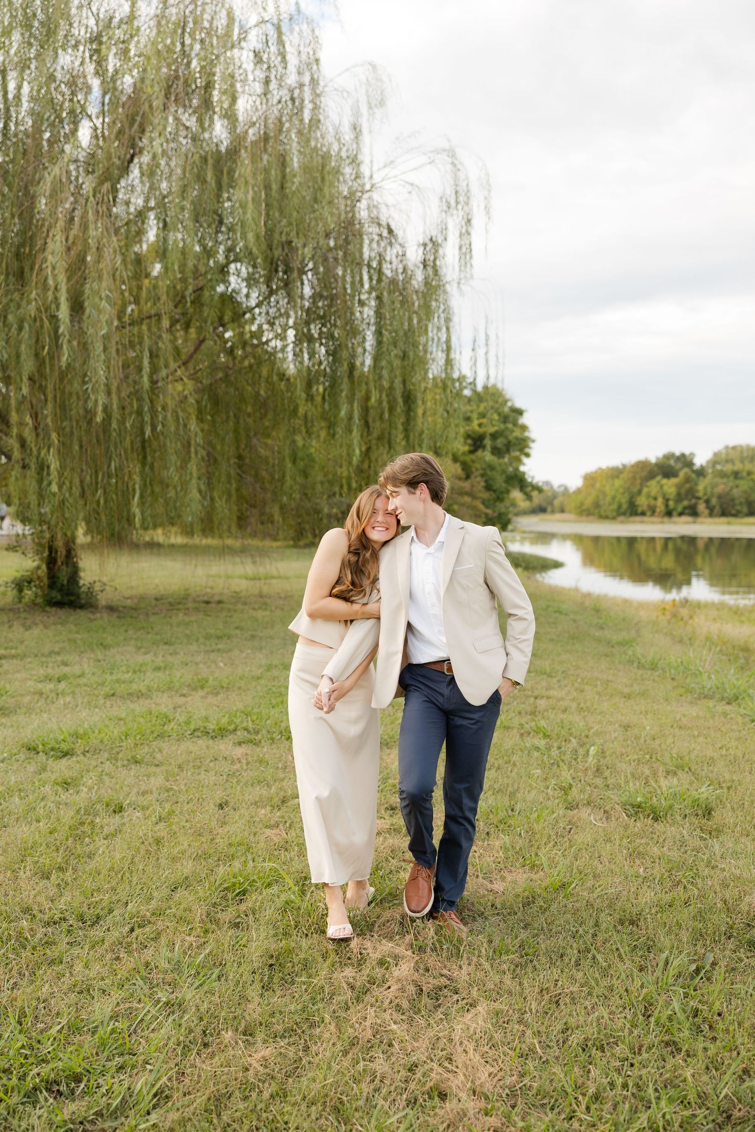 Romantic couple strolling along a grassy path beneath weeping willow trees on an overcast day.