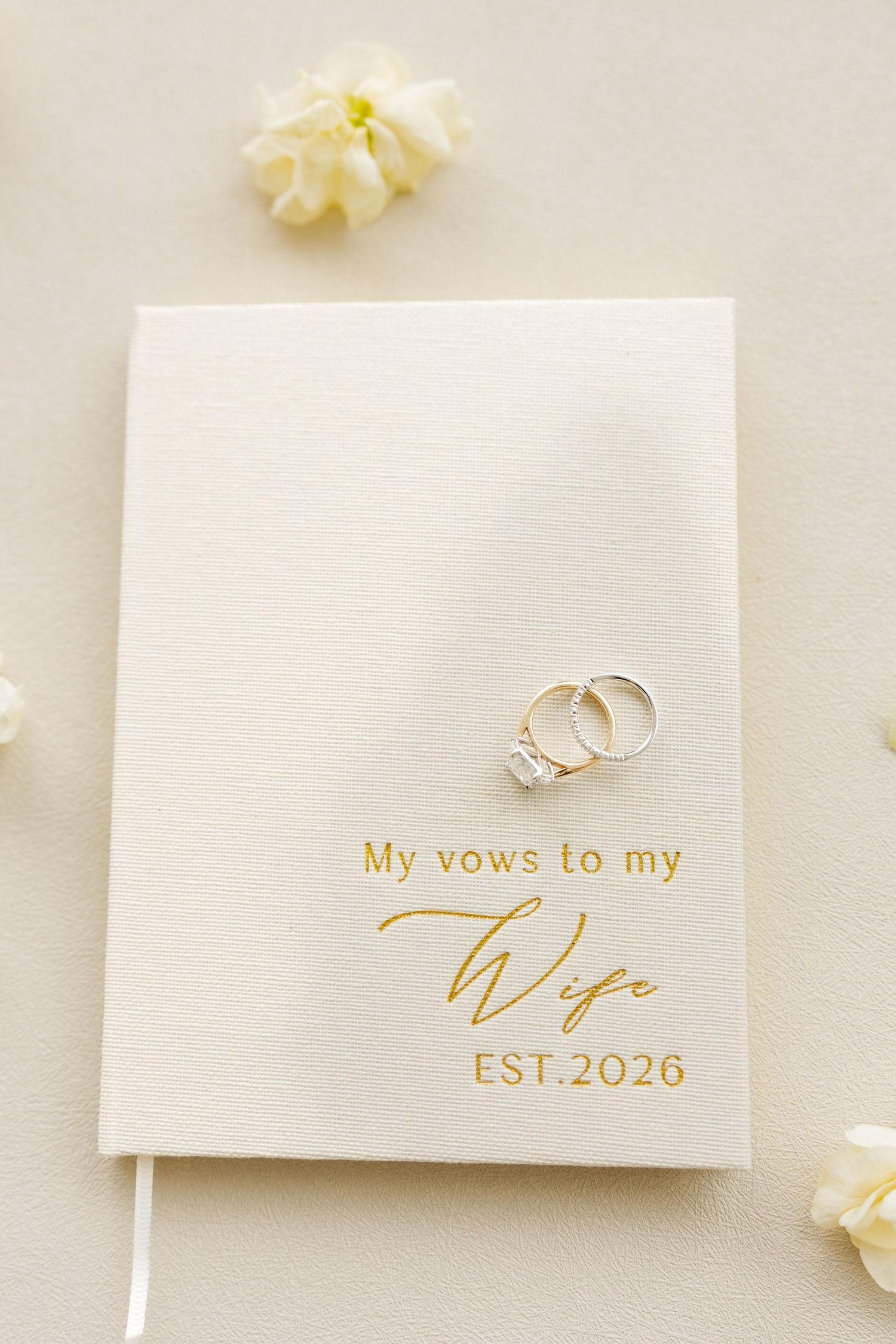 White vow book engraved with My Vows to my Wife EST.2026 in gold, topped with diamond engagement and wedding rings.