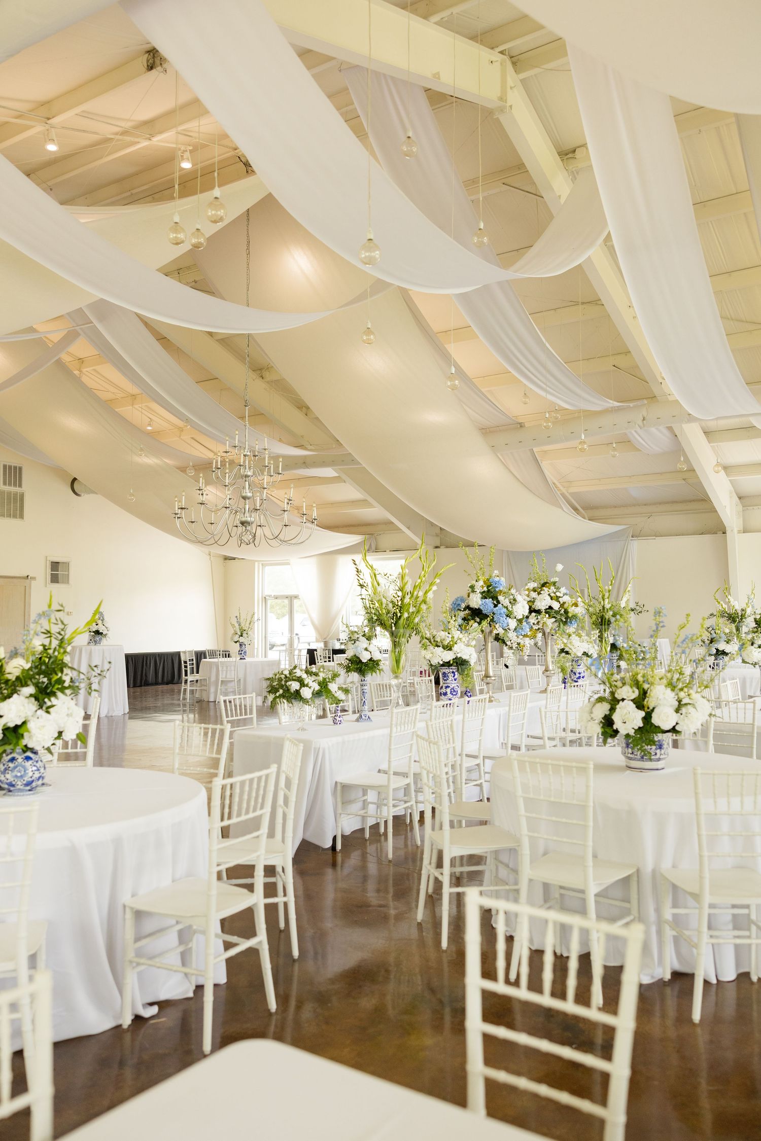 An elegant white reception hall with draped fabric ceiling, crystal chandeliers and floral centerpieces.Cold Springs Events.