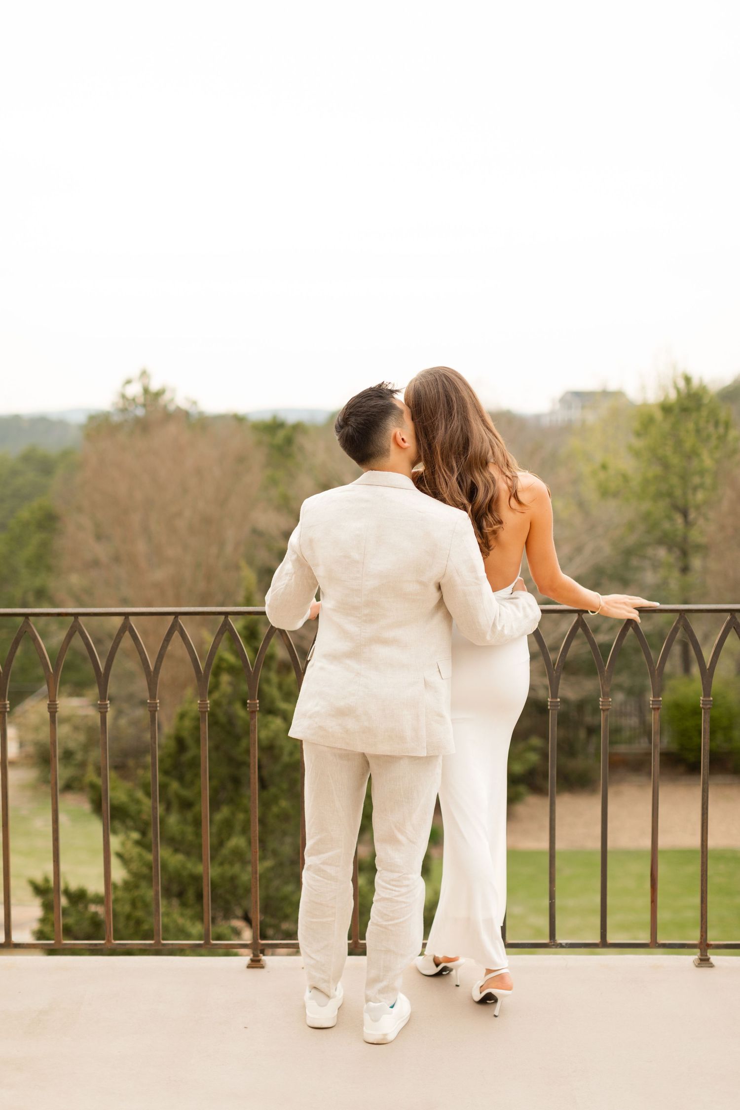 Two people in white outfits lean against an iron railing on a terrace overlooking a scenic landscape. Goodwin Manor.