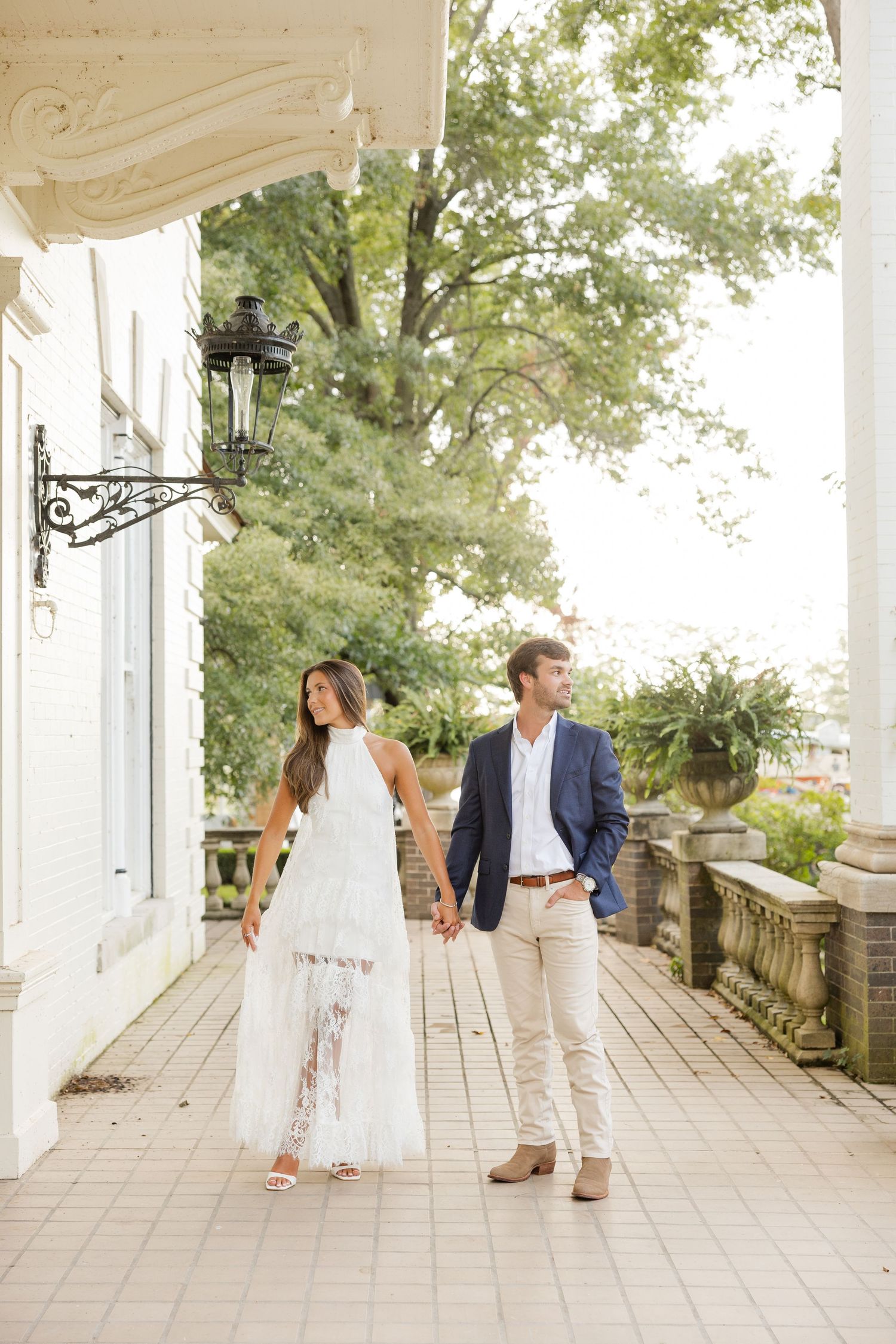 A couple strolls along a white columned porch during their engagement photo session.