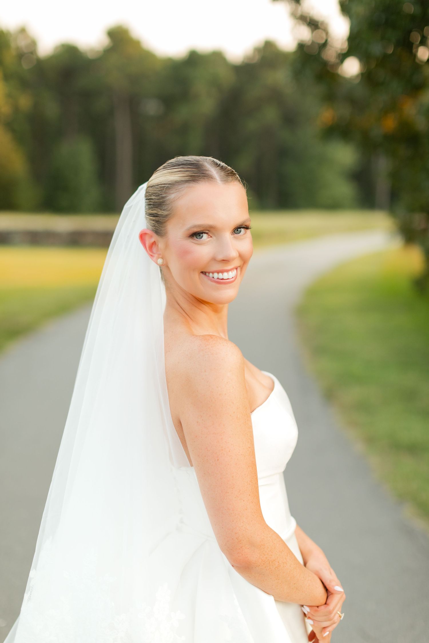 A bride in a white wedding dress and veil poses on a tree-lined path during golden hour.