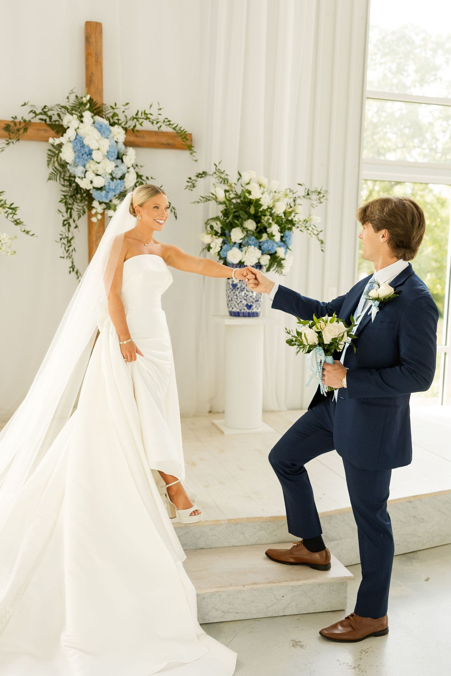 A playful moment between the couple on white chapel steps with floral arrangements.Cold Springs Events