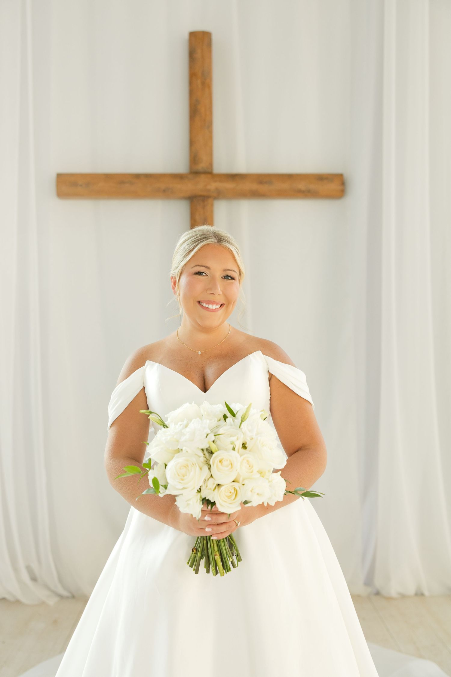 A bride in an off-shoulder white wedding gown holds a bouquet of white roses while standing beneath a wooden cross.