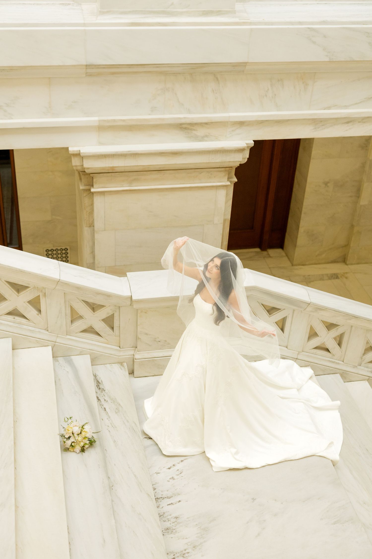 An elegant bridal gown with a flowing train cascades down marble steps in a classical setting.
