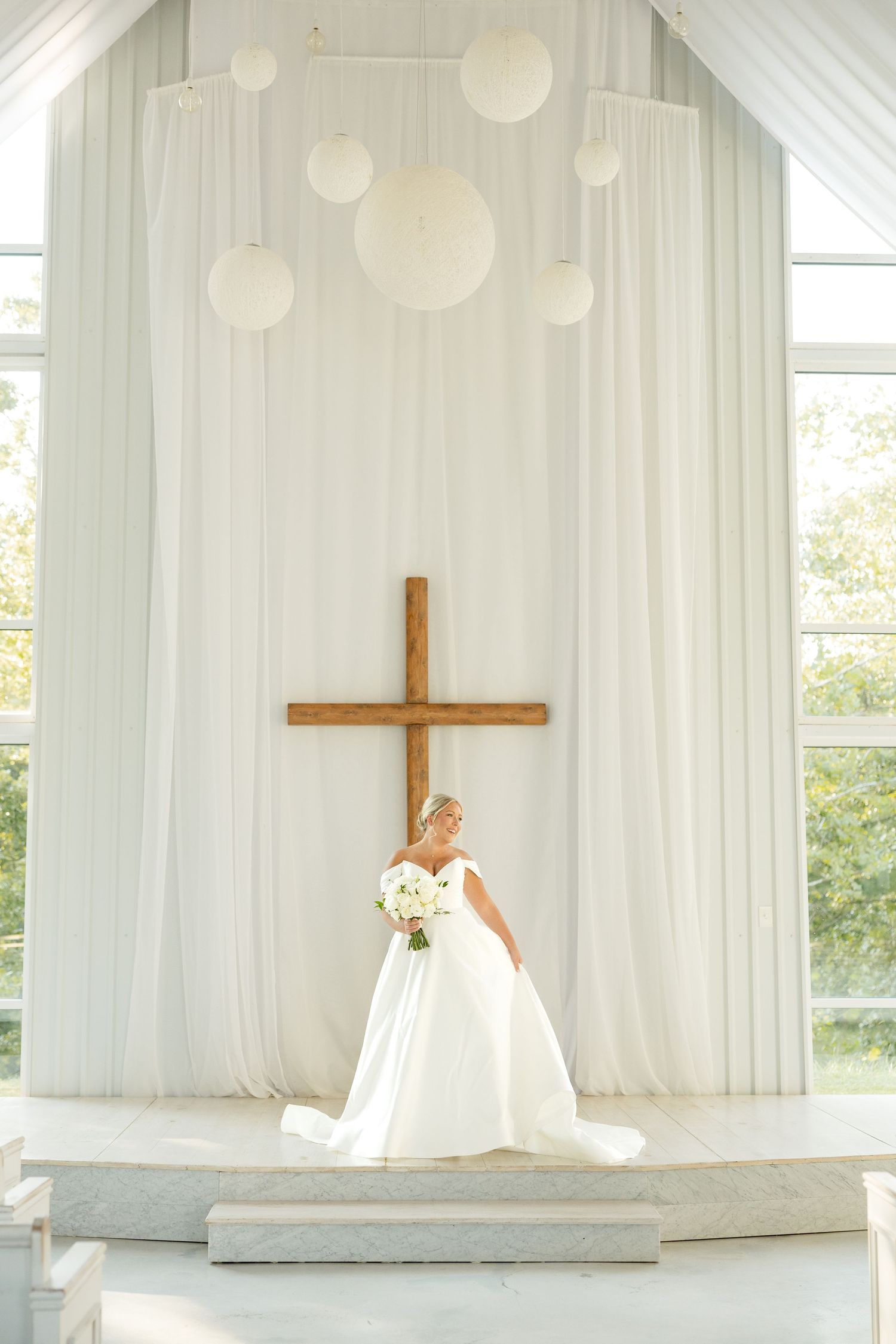 A bride in a white wedding dress poses in front of a cross in an elegant white chapel with natural lighting.