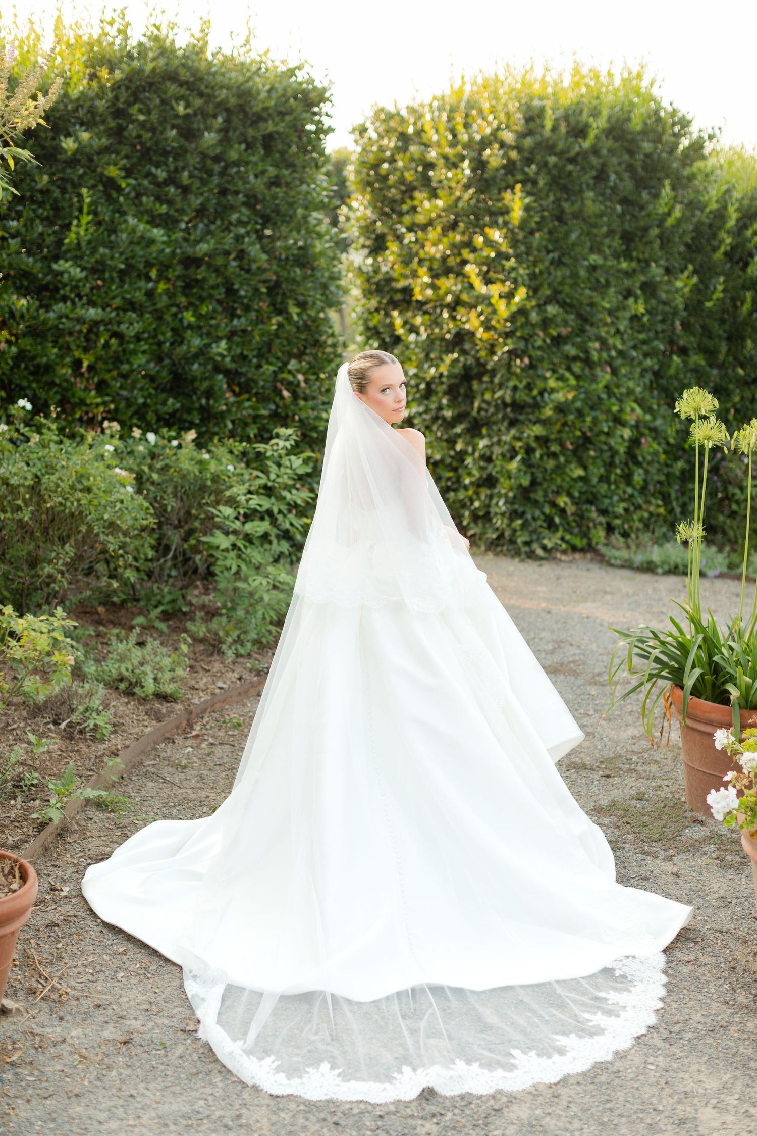 The train of a white wedding gown spreads gracefully across a garden path with lush greenery in the background.