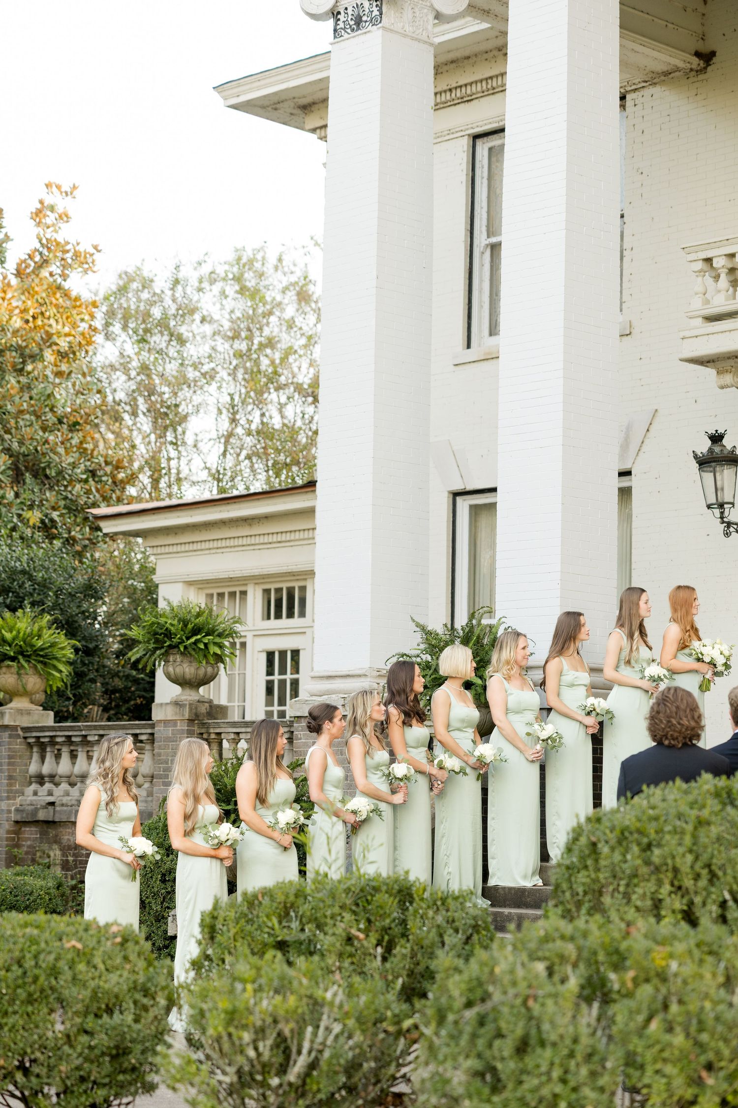Bridesmaids in sage green dresses stand in a row on white mansion steps holding bouquets during an elegant wedding.