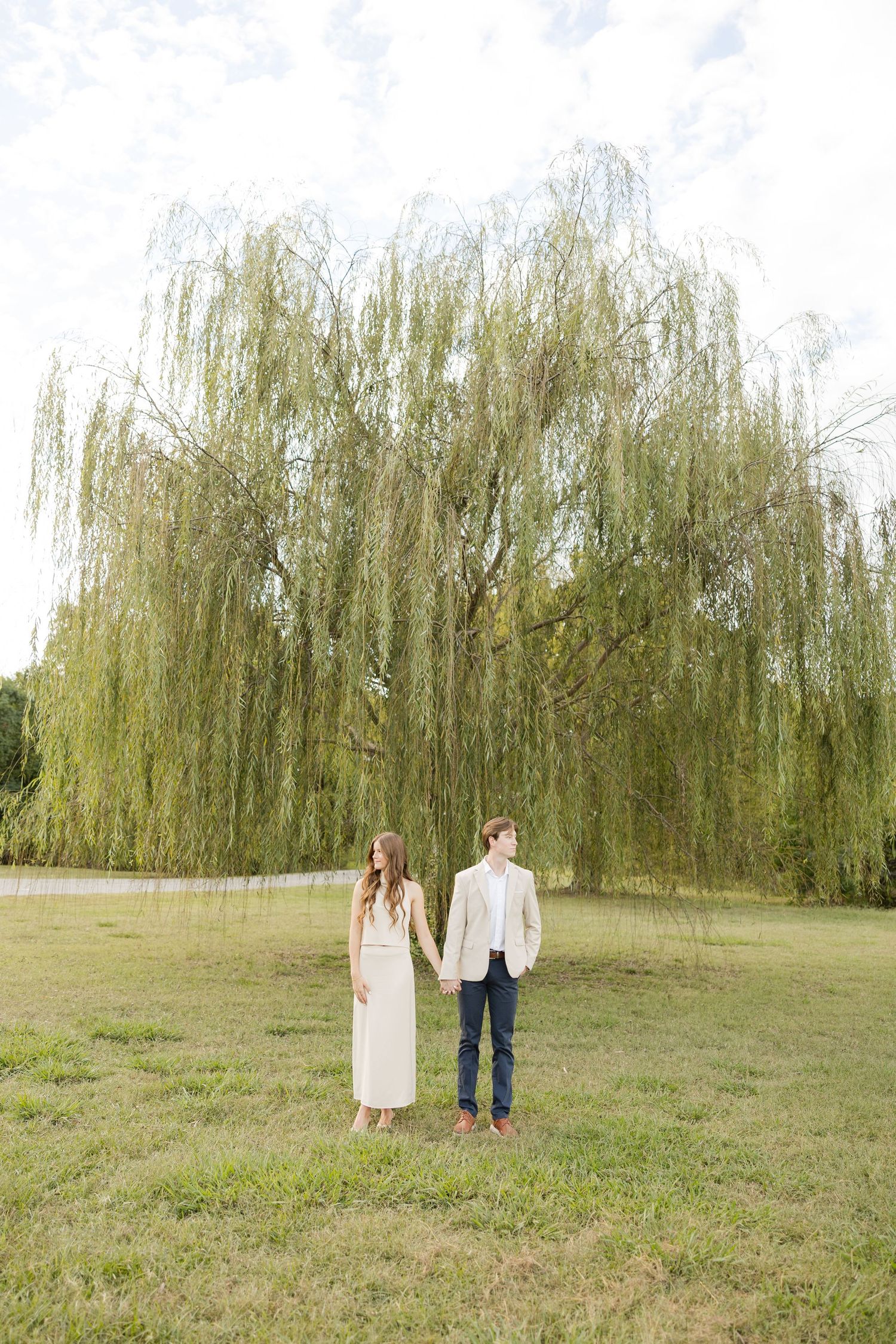 A couple stands beneath a majestic weeping willow tree in a serene park setting.