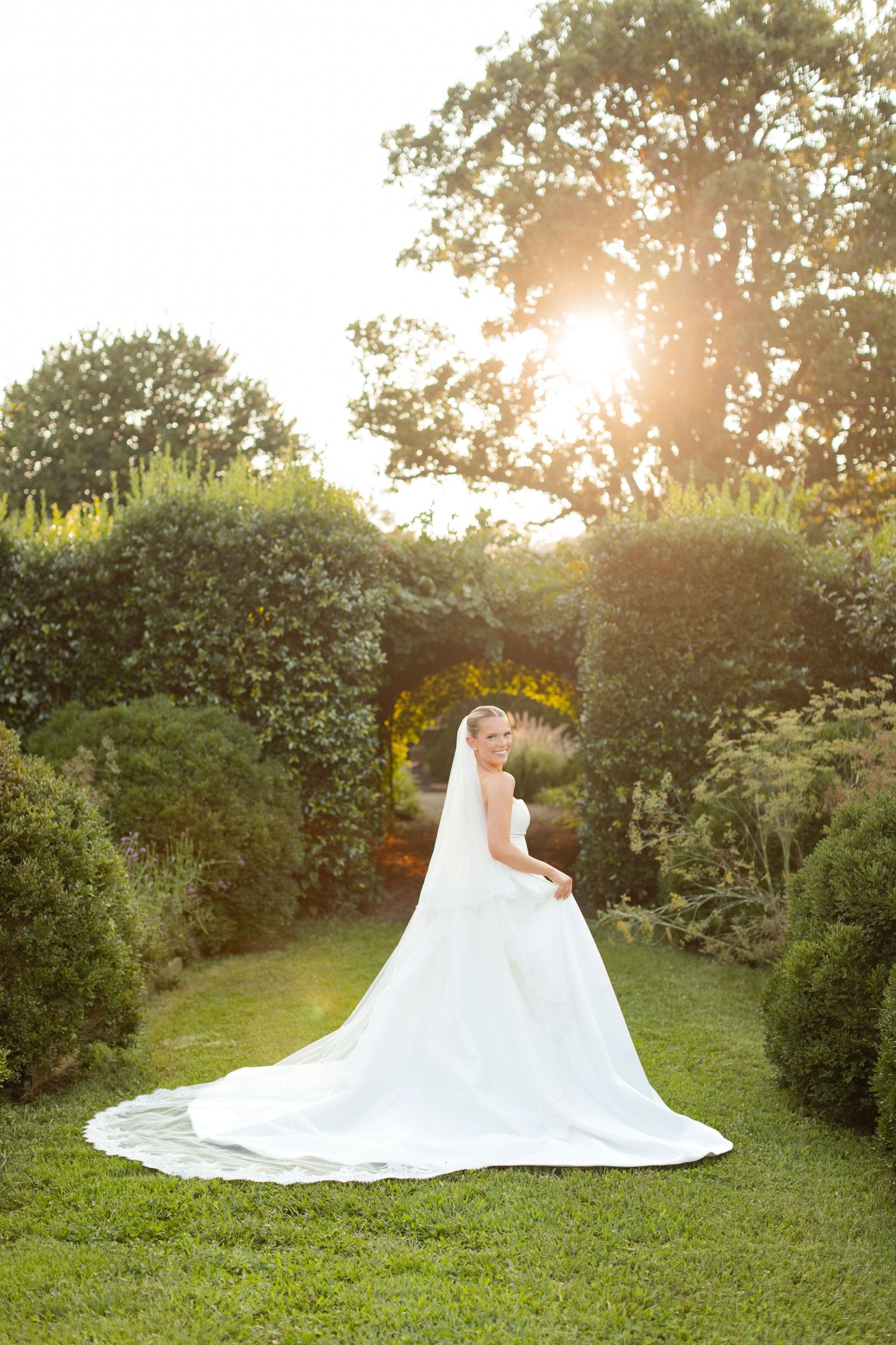 Elegant white wedding dress with flowing train photographed in a lush garden during golden hour.