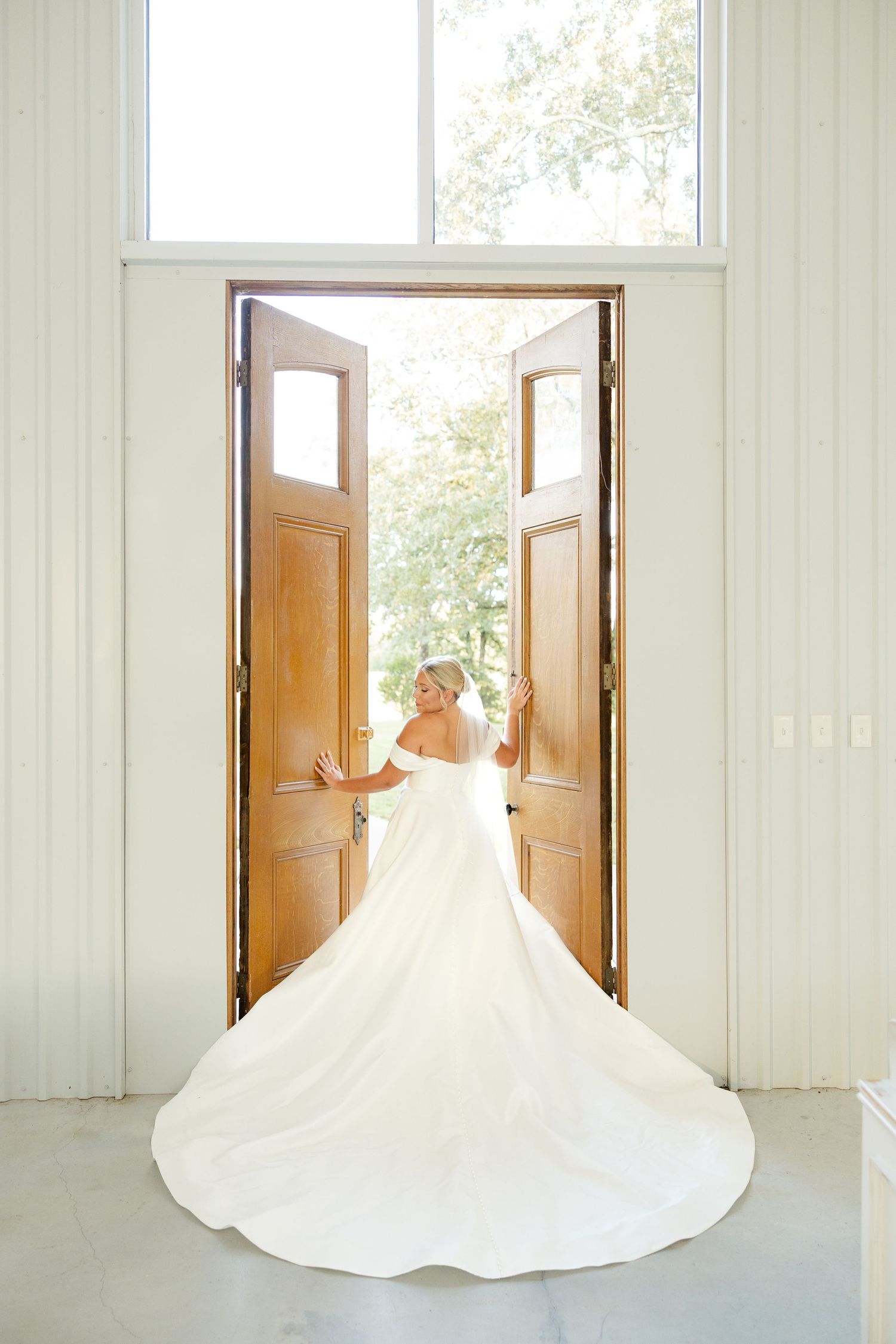 An elegant white wedding dress with a flowing train is photographed in a bright doorway with wooden double doors.