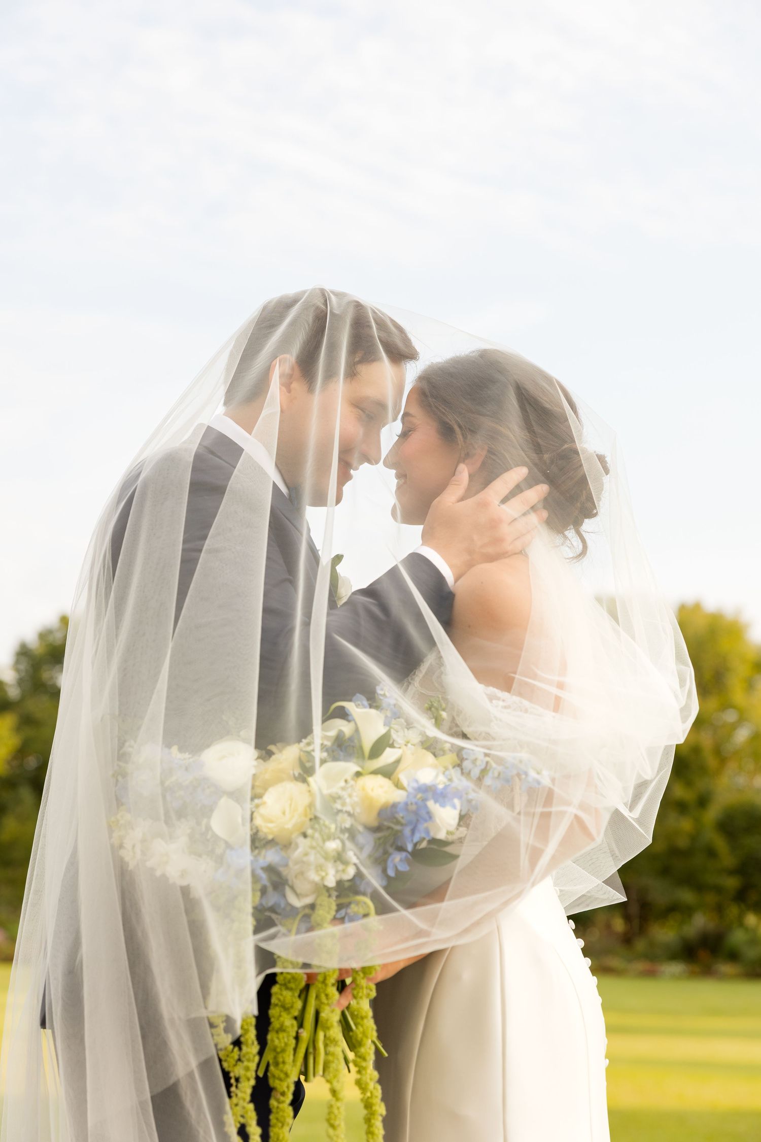 A romantic wedding couple shares an intimate moment under a flowing white veil in a sunlit outdoor garden setting.