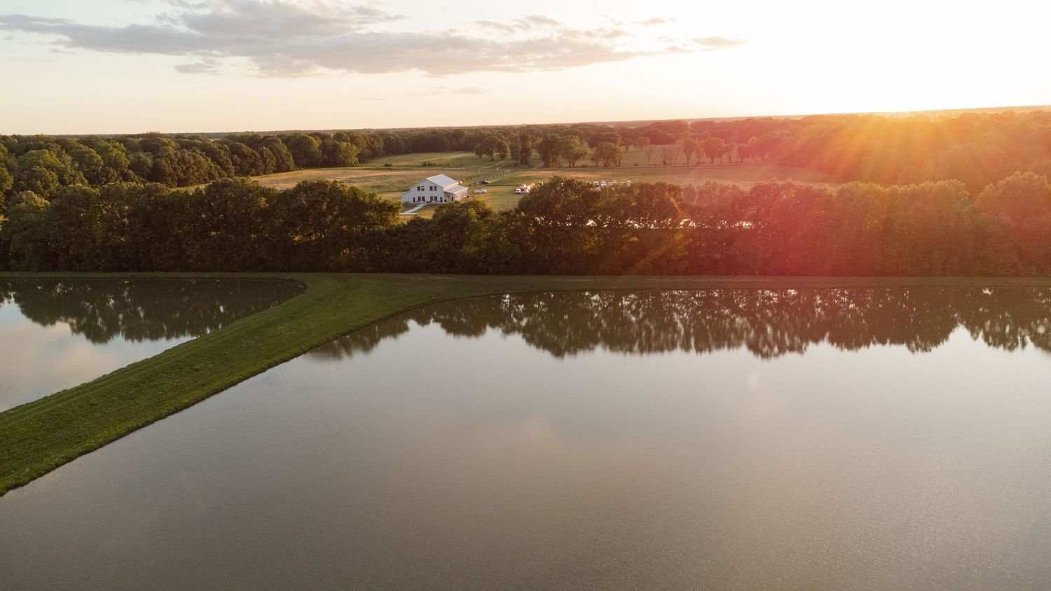 Aerial view of a tranquil lake reflecting golden sunset light with tree-lined shores and a path along the water's edge.