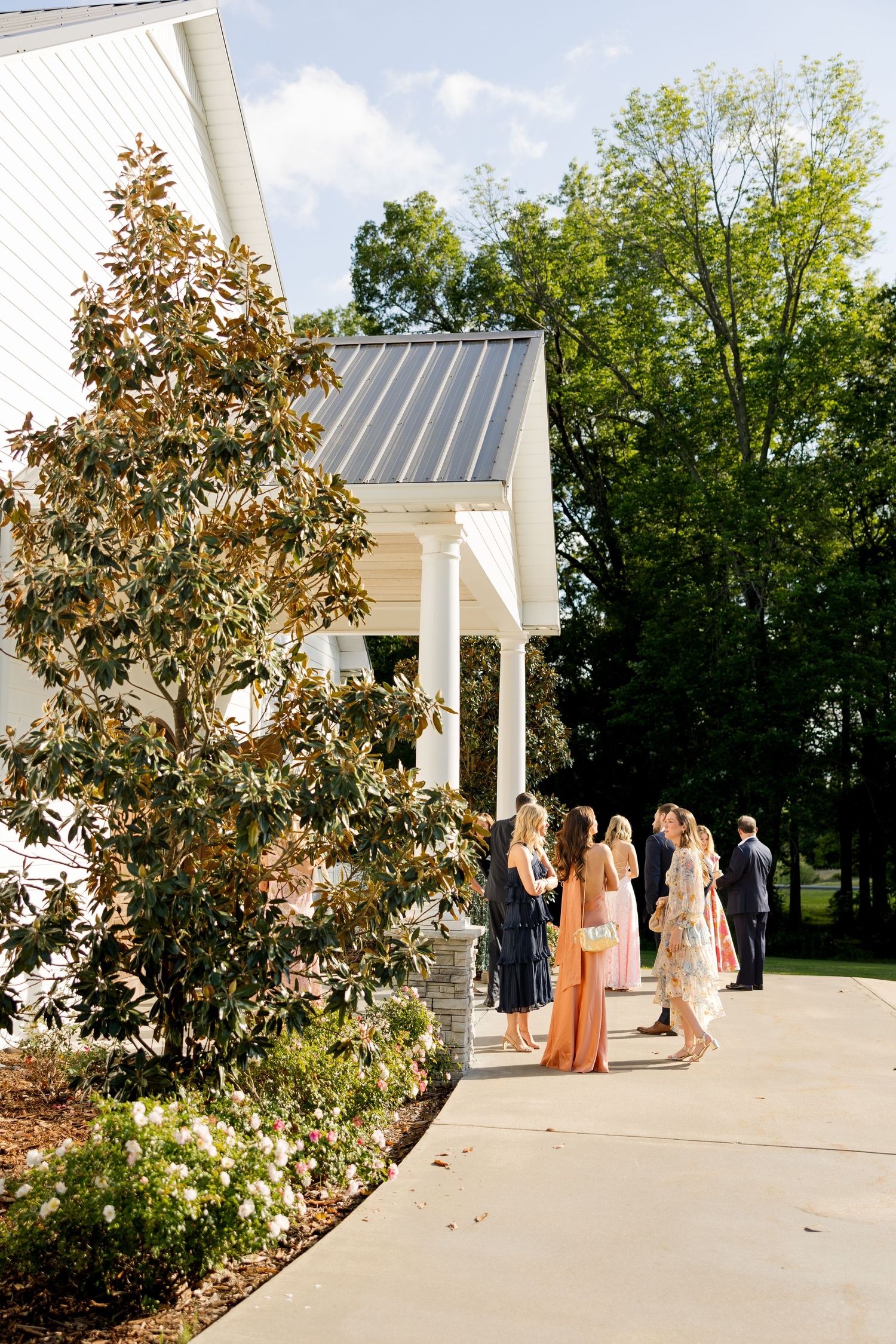 Guests in formal attire gather outside a white chapel with columns and metal roof, surrounded by lush greenery.