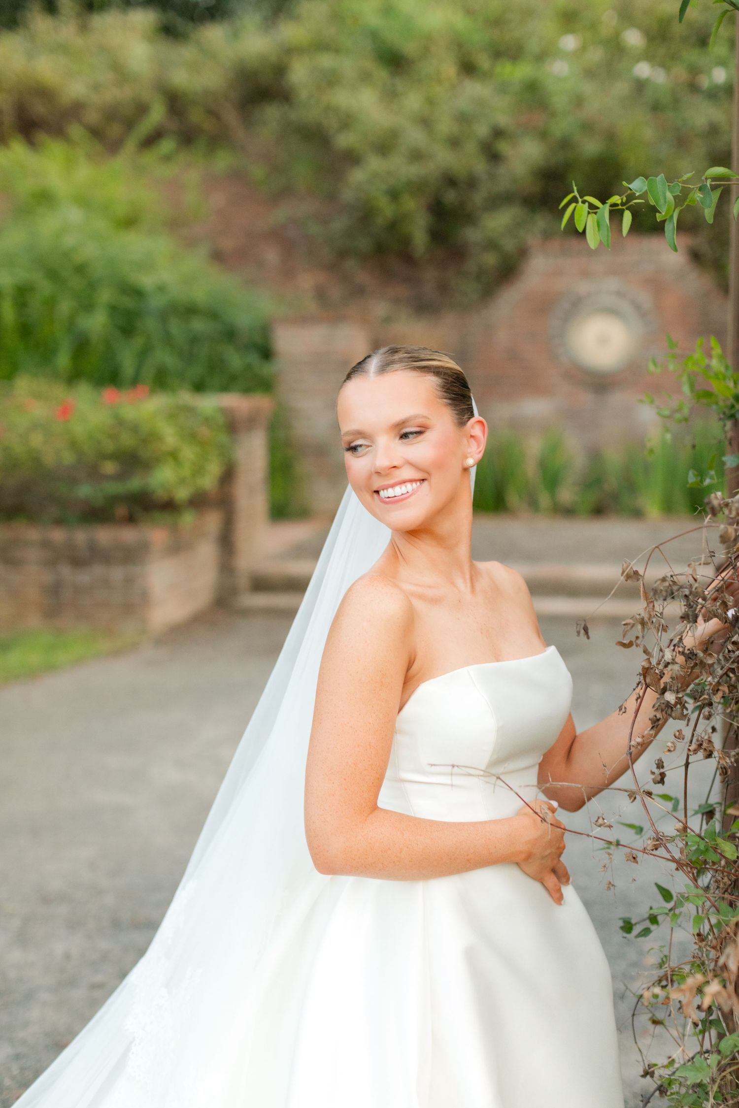 A bride in a strapless white gown poses gracefully in a garden setting with brick walls.