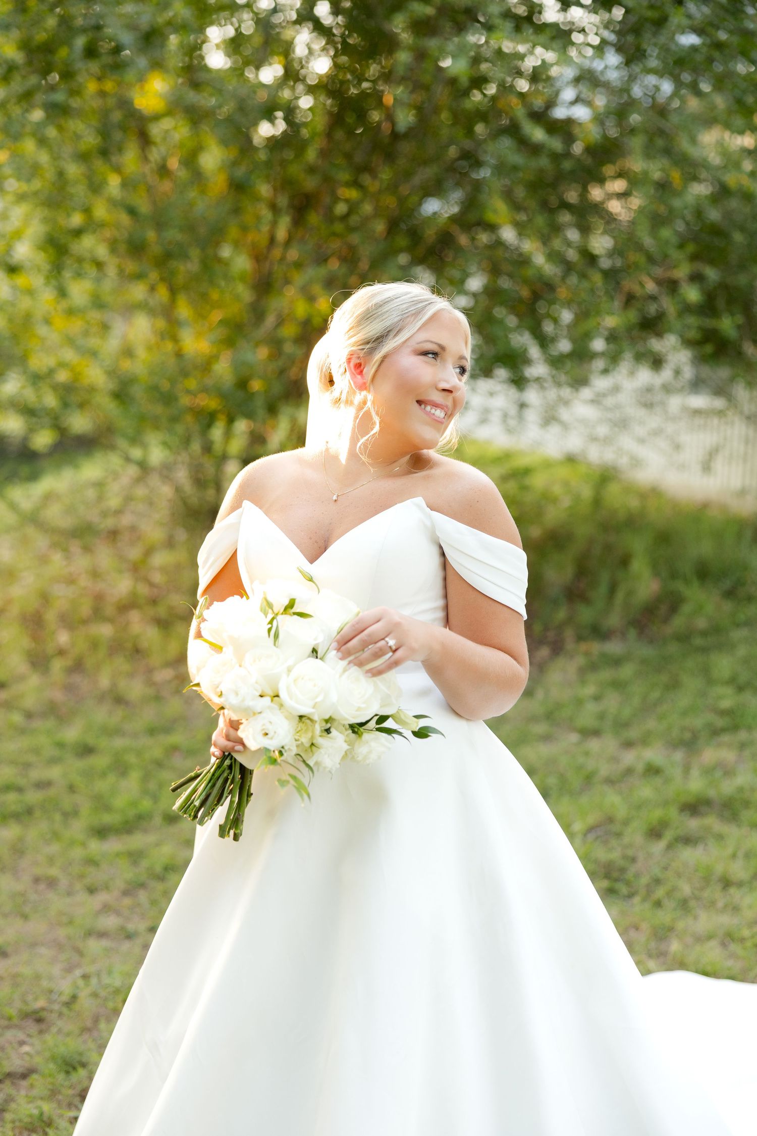A bride in an elegant off-shoulder wedding gown twirls joyfully in a sunlit garden setting.