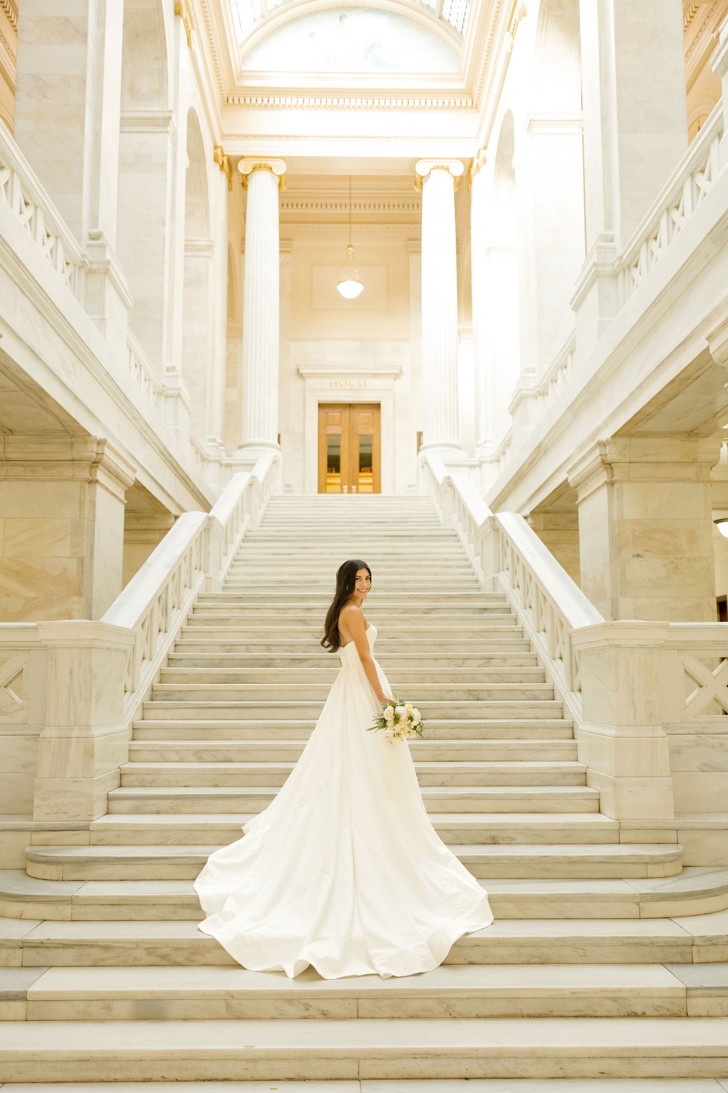 Elegant wedding dress with flowing train photographed on grand marble staircase in classical building interior.