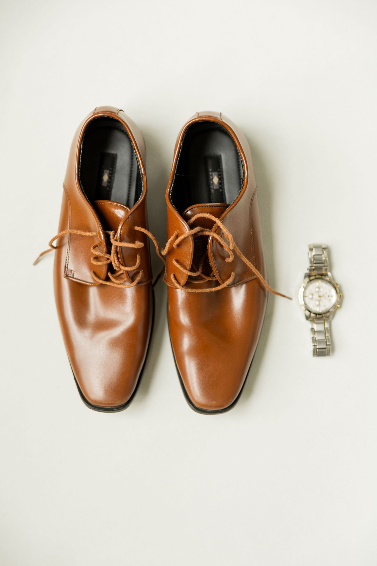 Brown leather dress shoes and silver watch arranged on white background for wedding details.Cold Springs Events