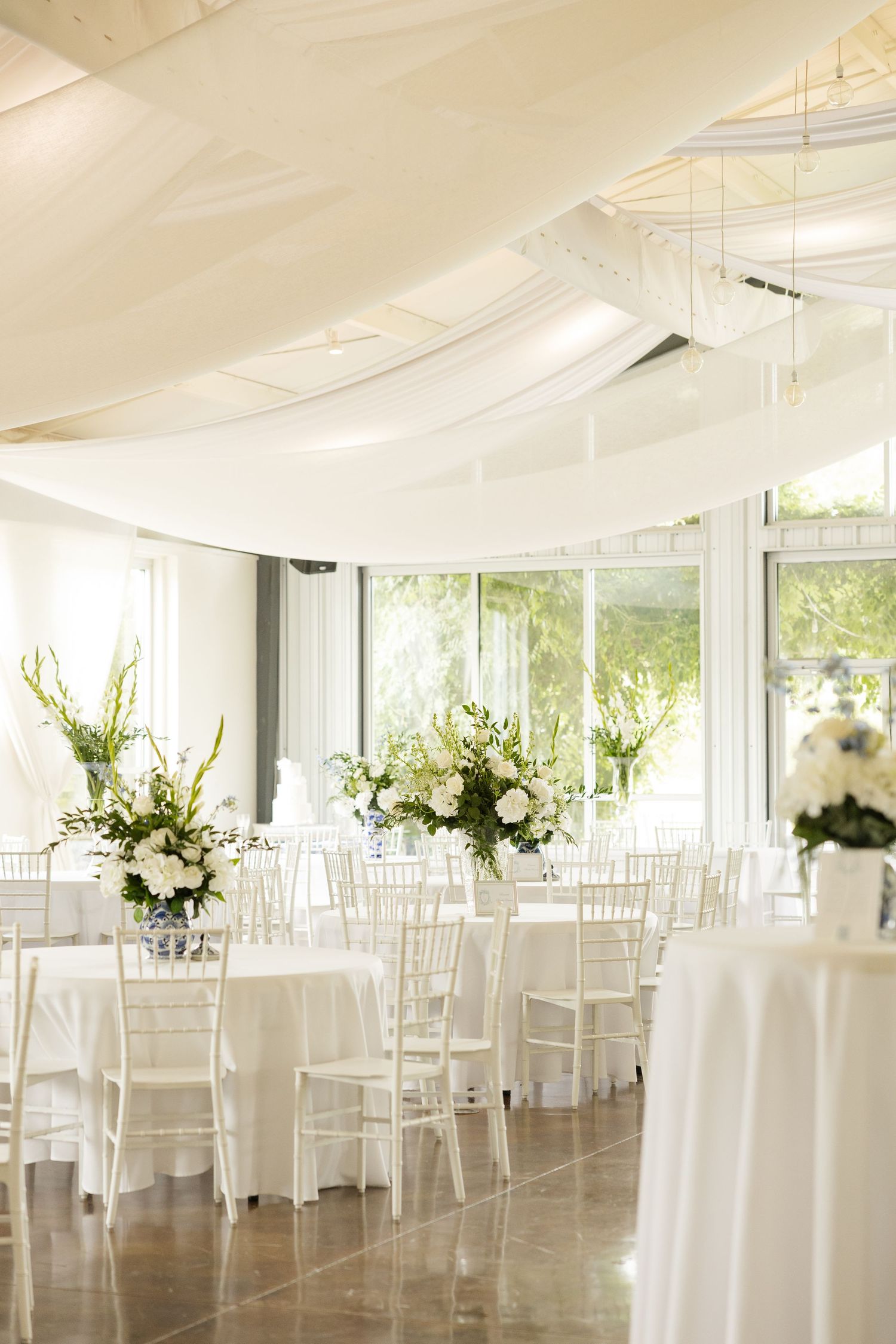 Elegant white wedding reception venue with draped fabric ceiling, white chiavari chairs, and tall floral centerpieces.Cold Springs Events