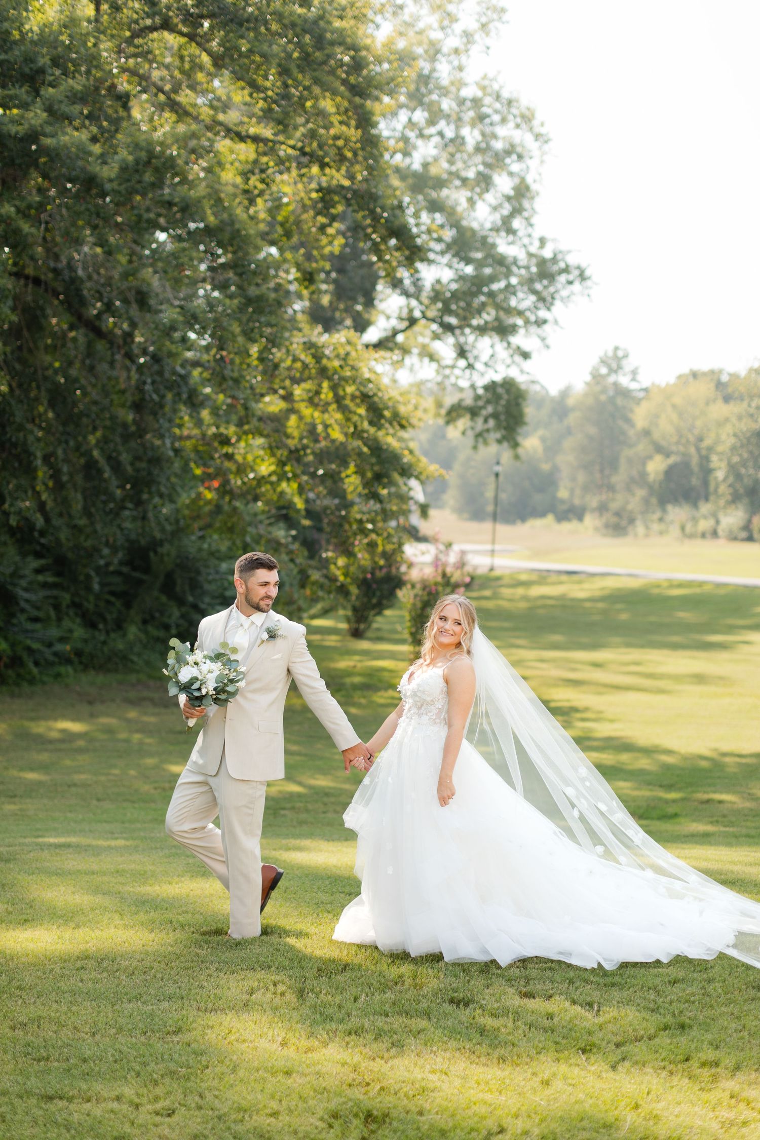 A couple walks hand in hand across a sunlit lawn in wedding attire. Legacy Acres.