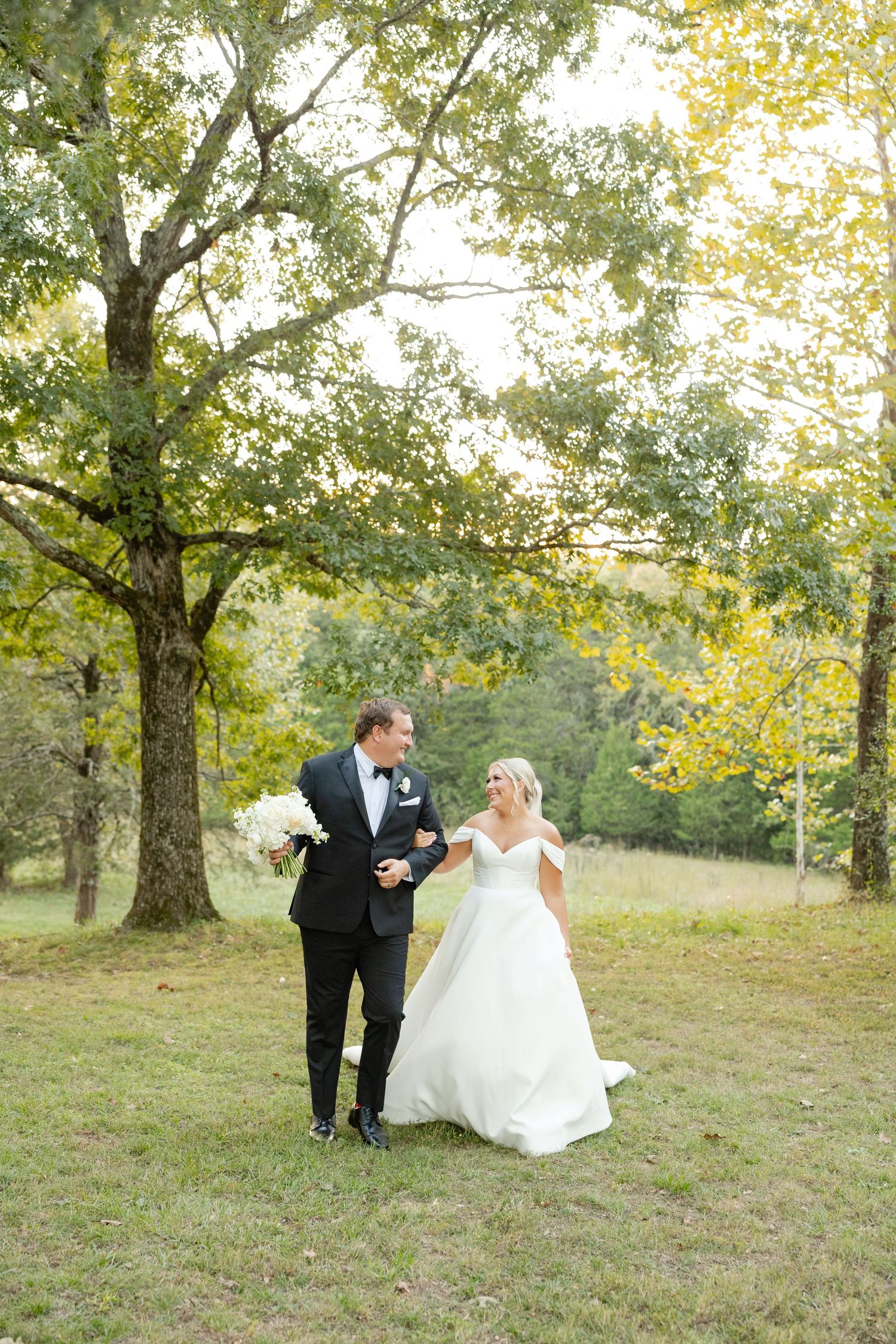A couple in formal wedding attire walks together through a scenic field with autumn trees in the background. Cold Springs Events.