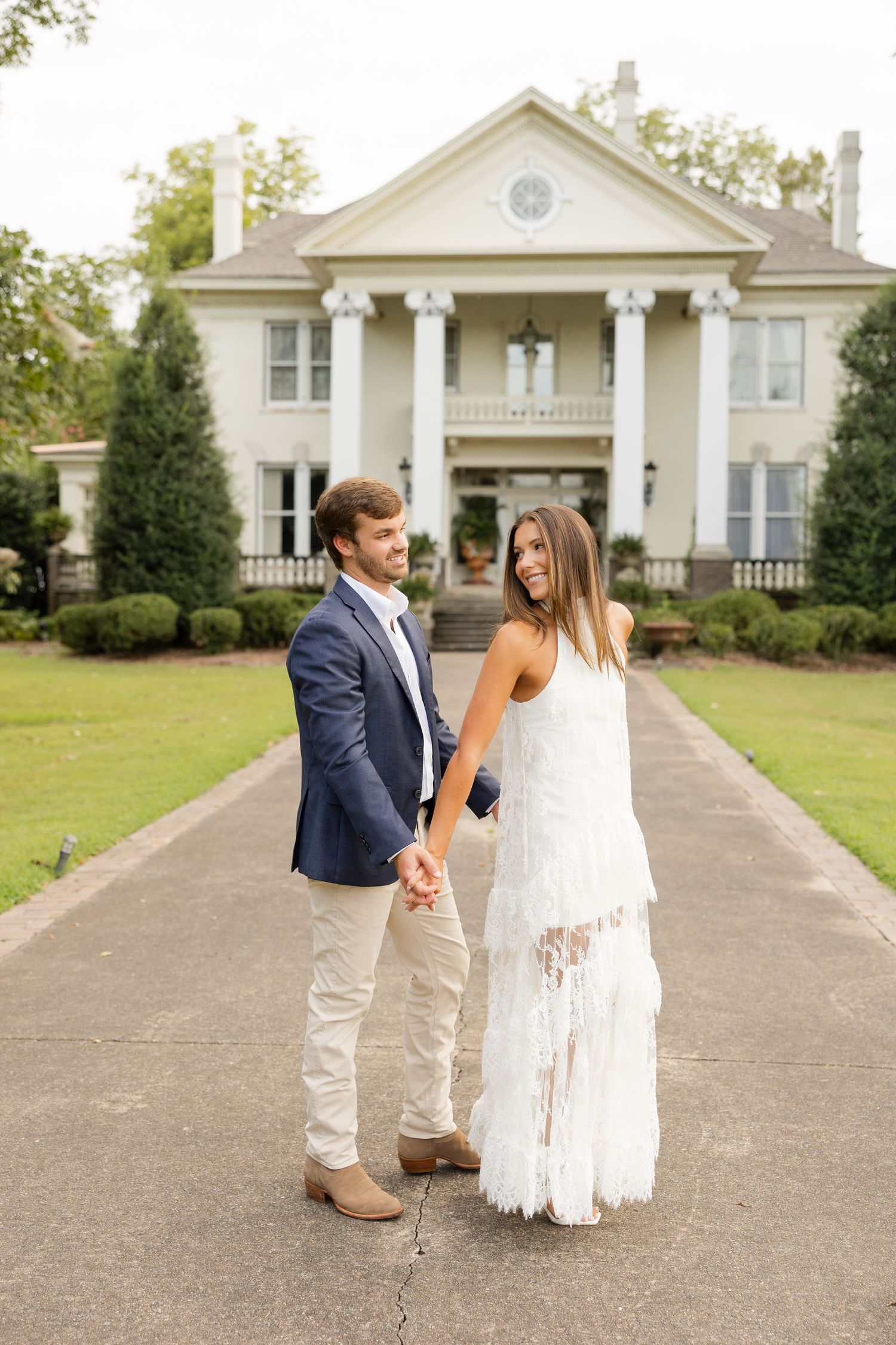 Couple in wedding attire walking and posing playfully on a path in front of a white mansion.