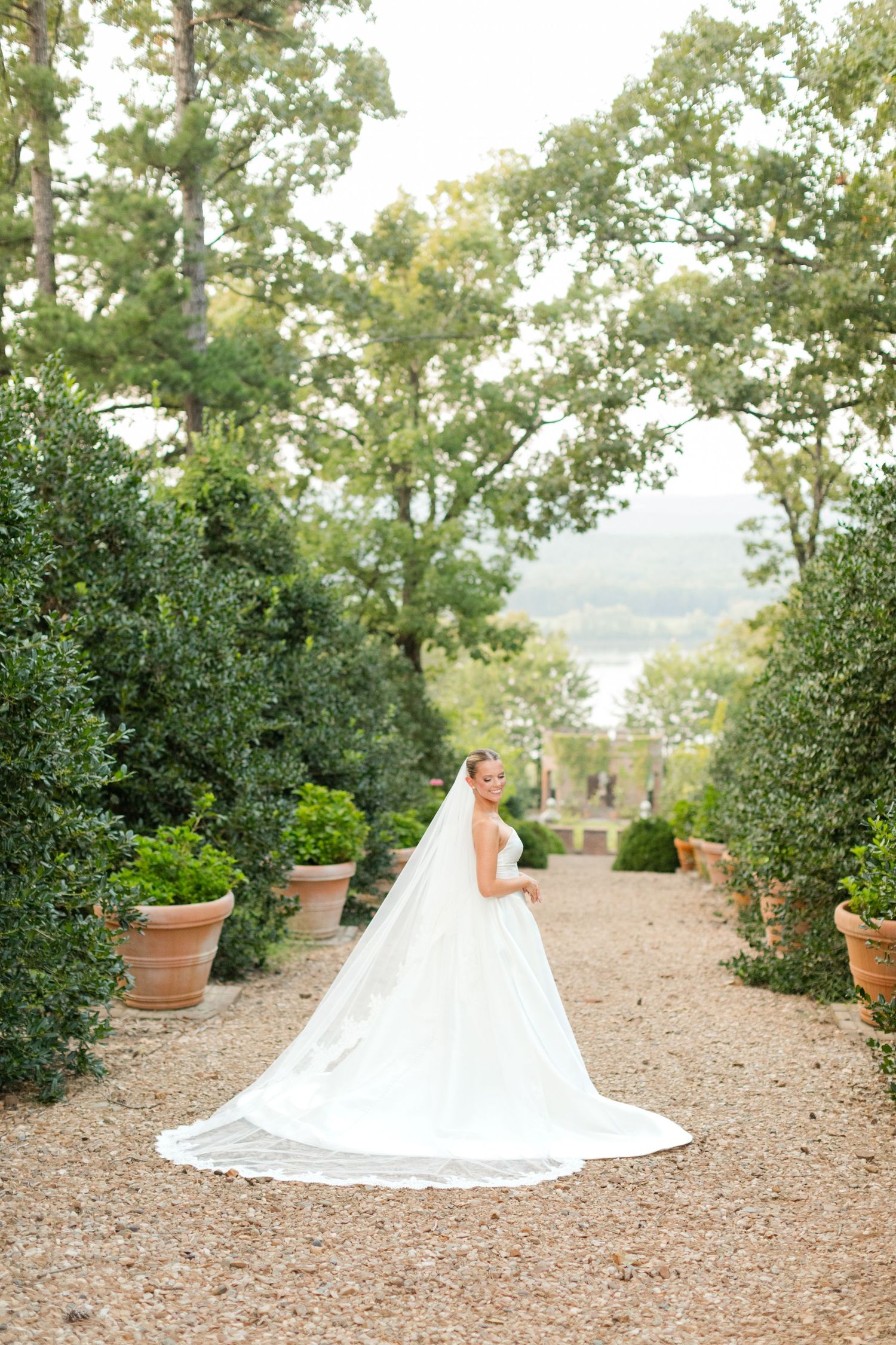 An elegant white bridal gown trails along a garden path surrounded by potted plants and greenery.